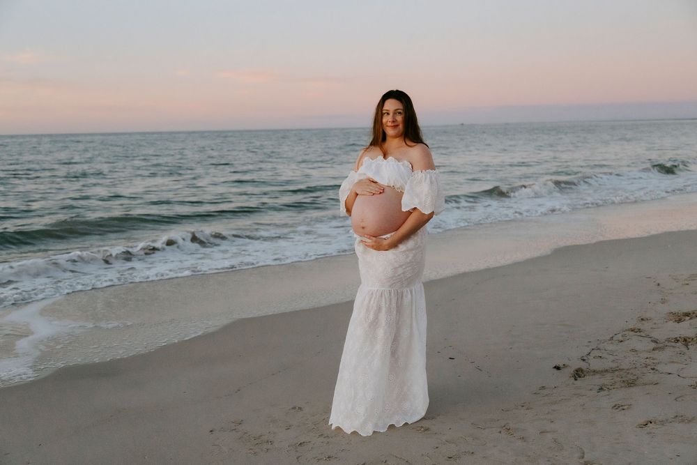 Pregnant woman in white off-shoulder lace gown poses on sandy beach at sunset with ocean waves behind her.