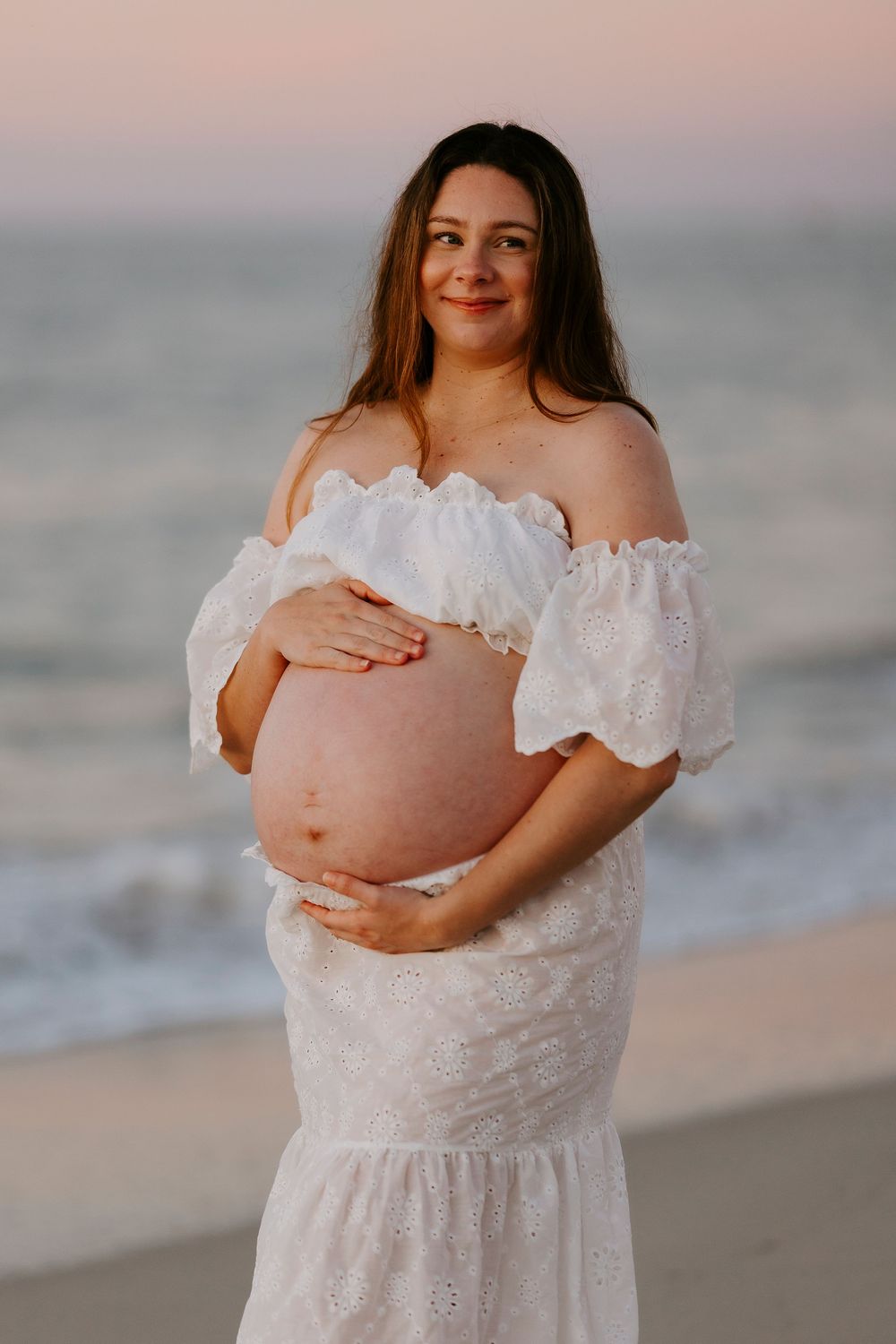 Pregnant woman in white off-shoulder dress cradling baby bump on beach at sunset.