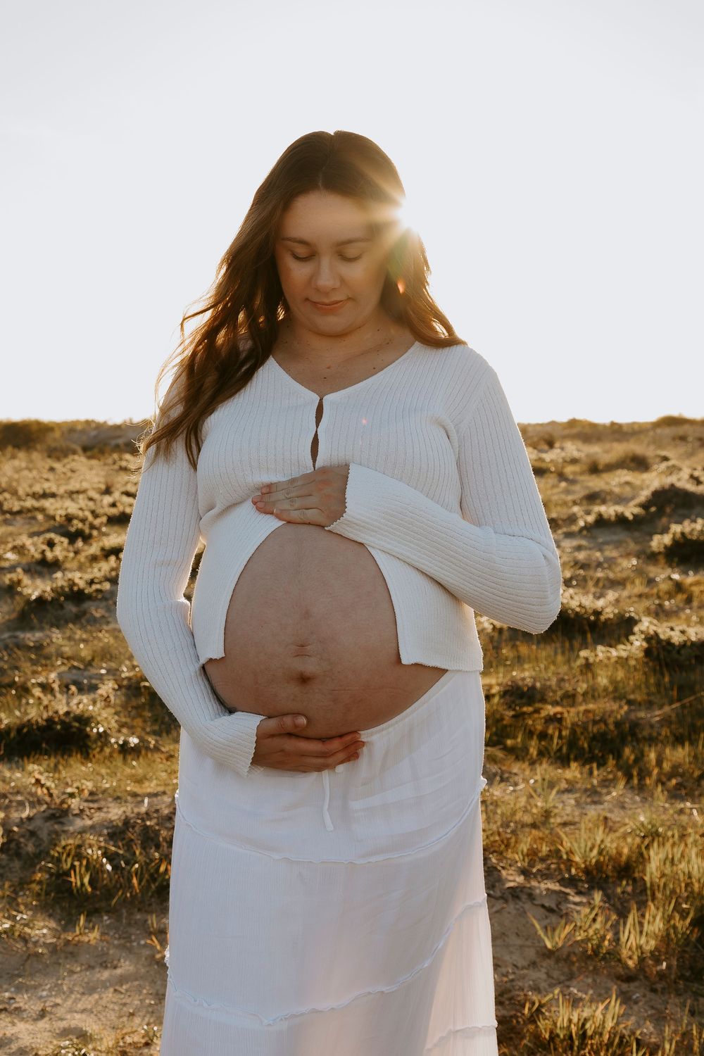 Pregnant woman in white dress holding baby bump outdoors at sunset in a serene desert landscape.