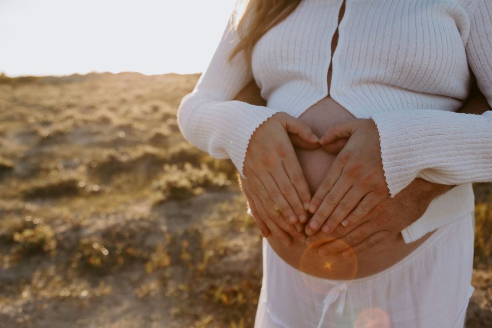 Pregnant woman in white dress holding baby bump with both hands in a sunny golden field.