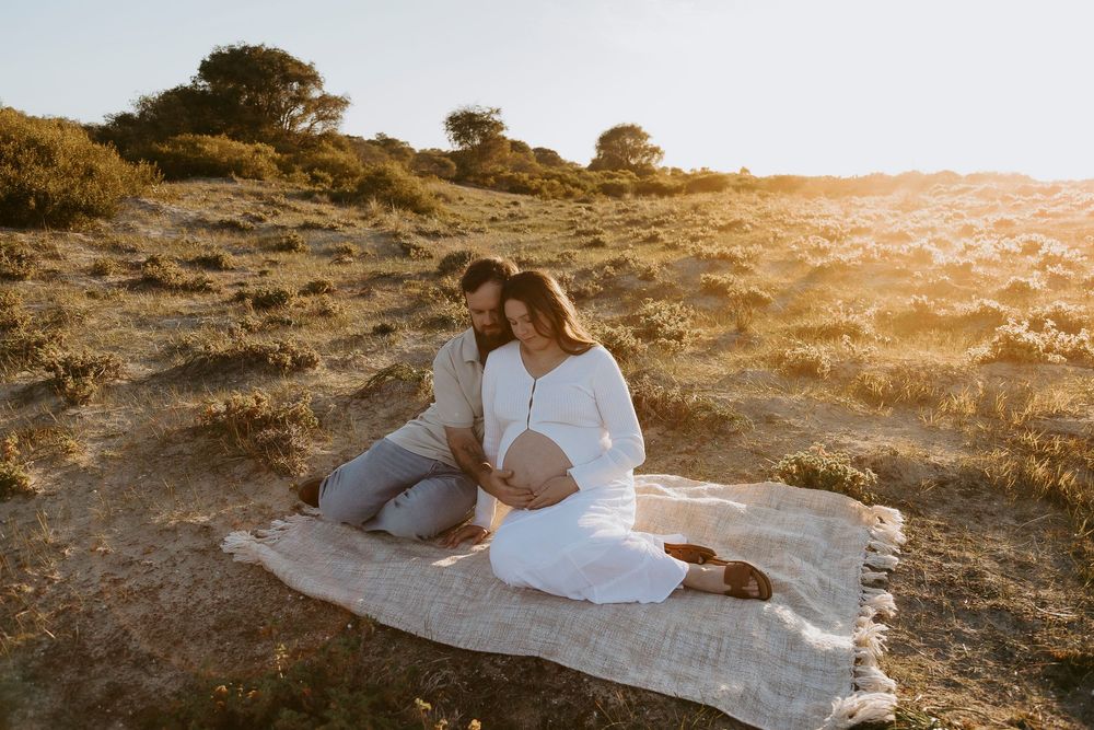 Couple sitting on blanket in golden field at sunset, woman in white dress with baby bump, man embracing her tenderly.