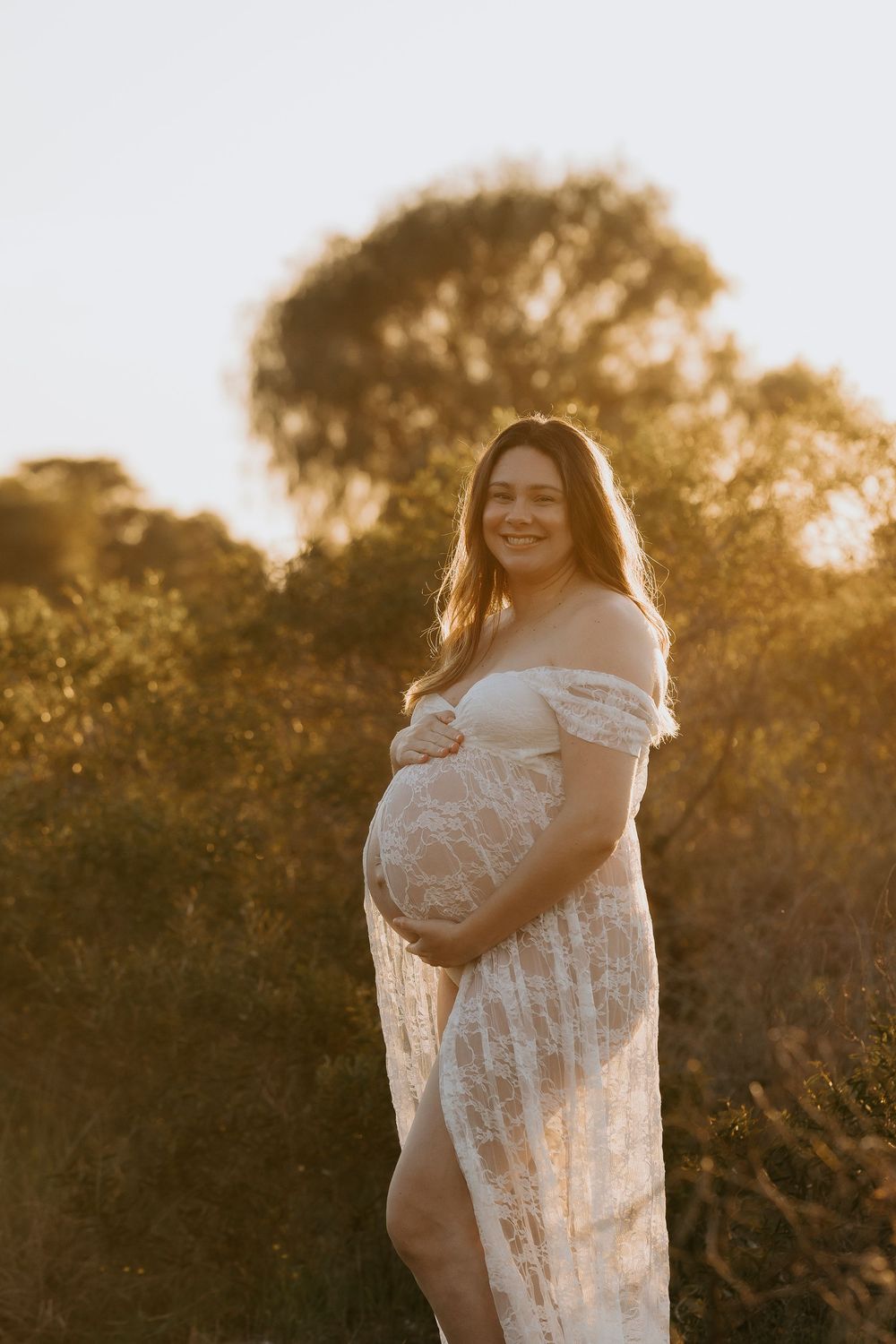 Smiling pregnant woman in white lace off-shoulder gown cradling her belly outdoors at golden hour.