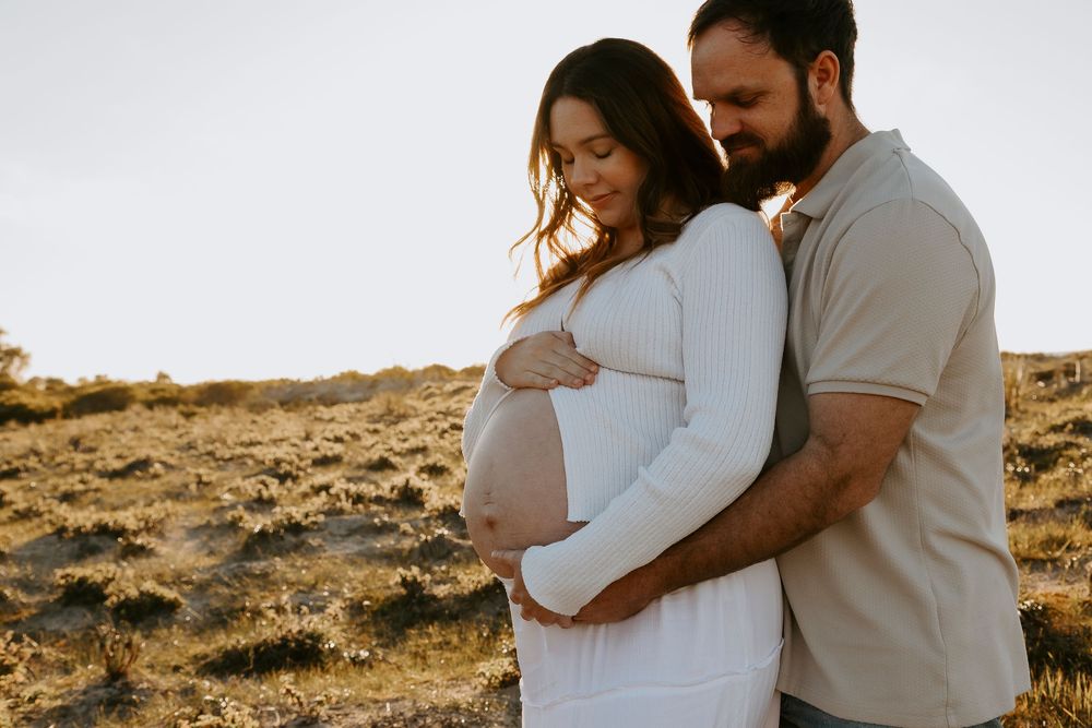 Couple embracing outdoors during golden hour maternity photoshoot, pregnant woman in white dress cradling baby bump.