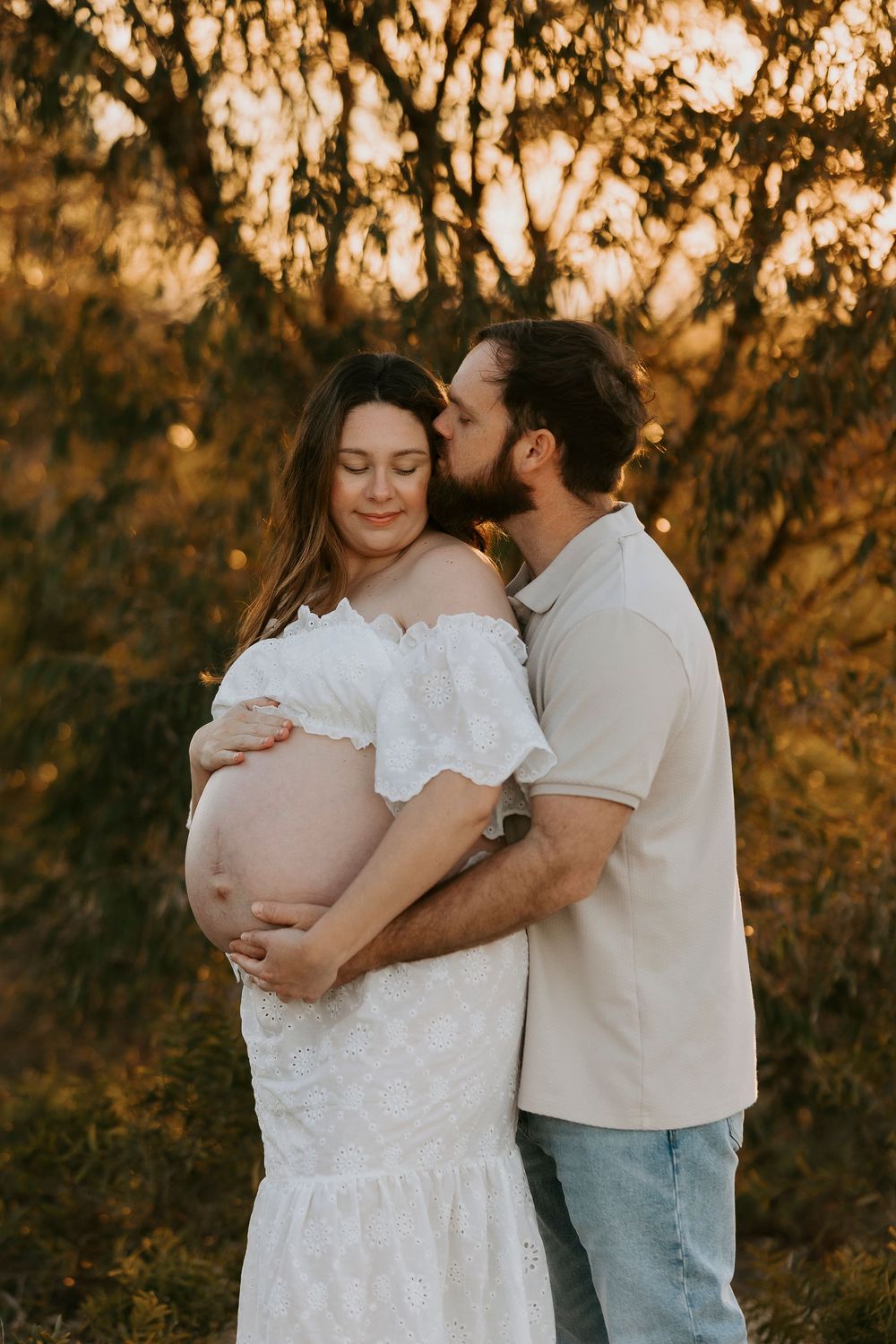 A couple shares a tender maternity moment at golden hour, man kissing pregnant woman in white dress outdoors.