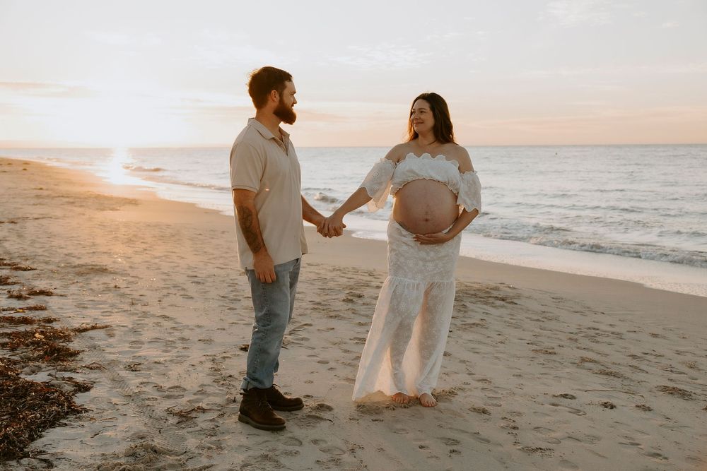 Pregnant couple holding hands on a sandy beach at sunset, woman in white lace maternity dress showing baby bump.