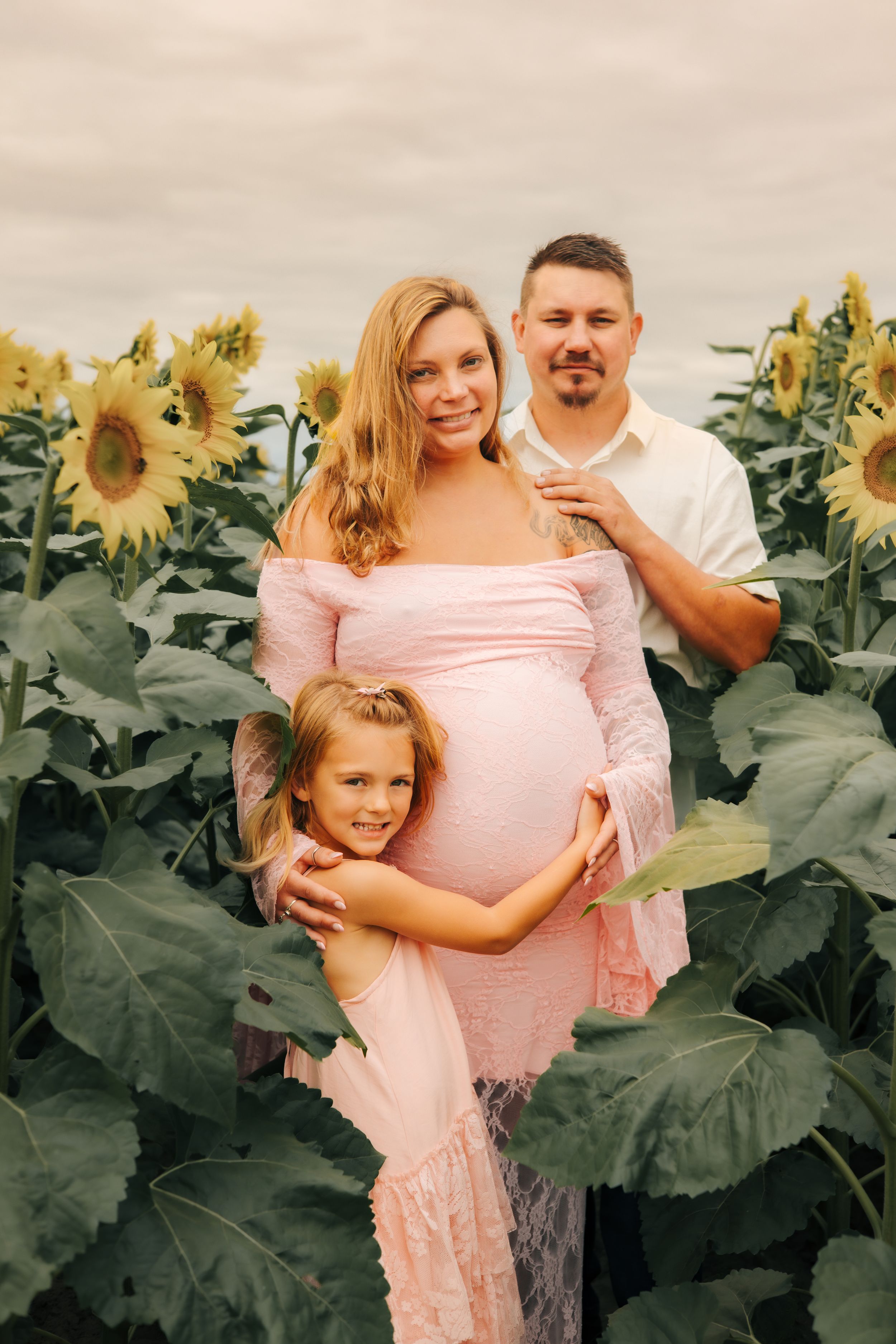 Pregnant woman in pink dress with husband and daughter in sunflower field for maternity photo shoot.