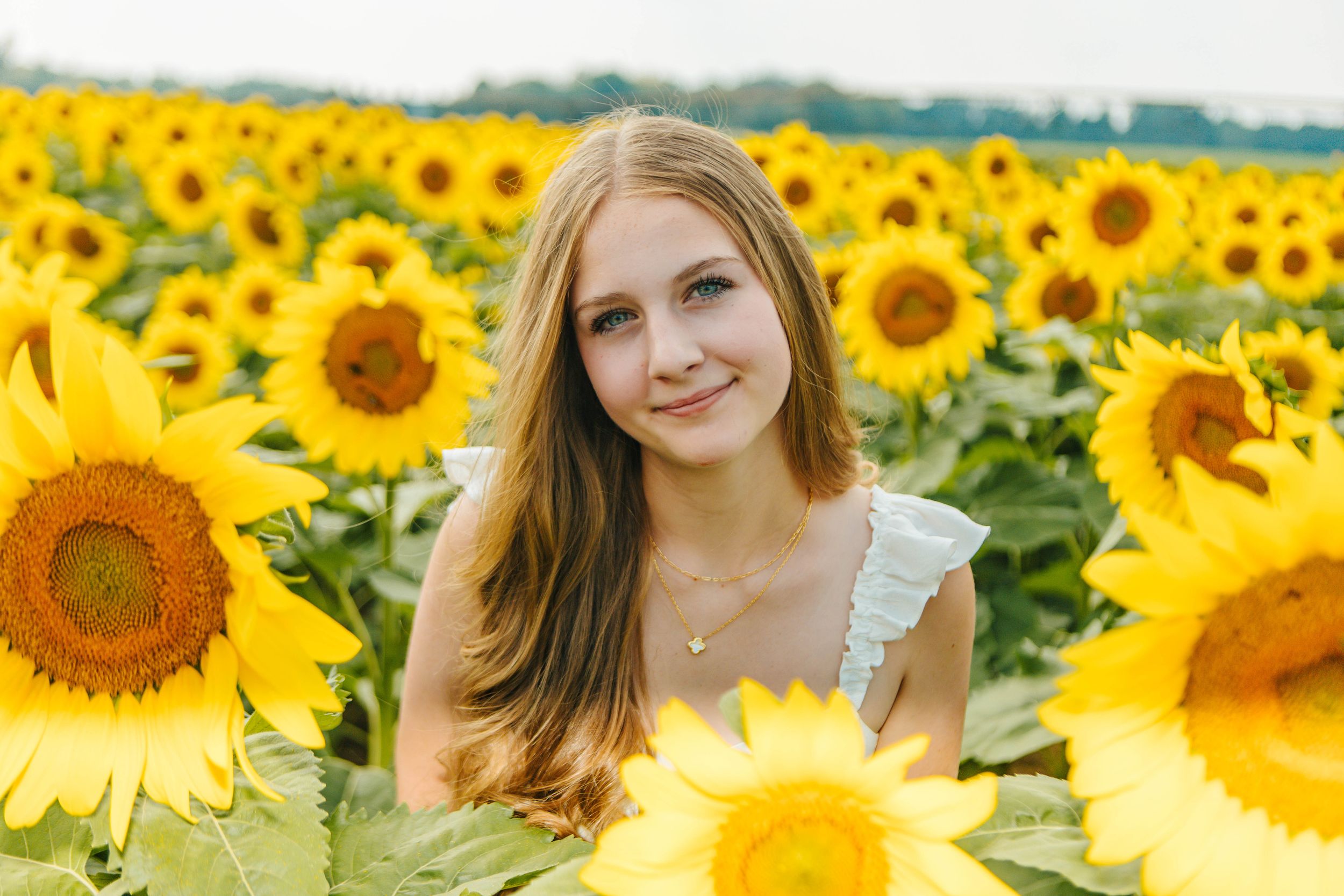 Young woman with long hair smiling in a vibrant sunflower field, wearing a white dress and gold necklace.