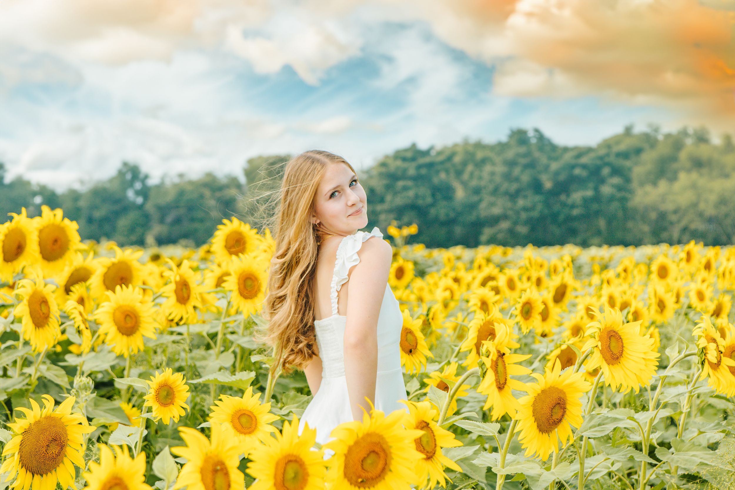 Young blonde woman in white dress standing in a vibrant sunflower field under a dramatic cloudy sky.