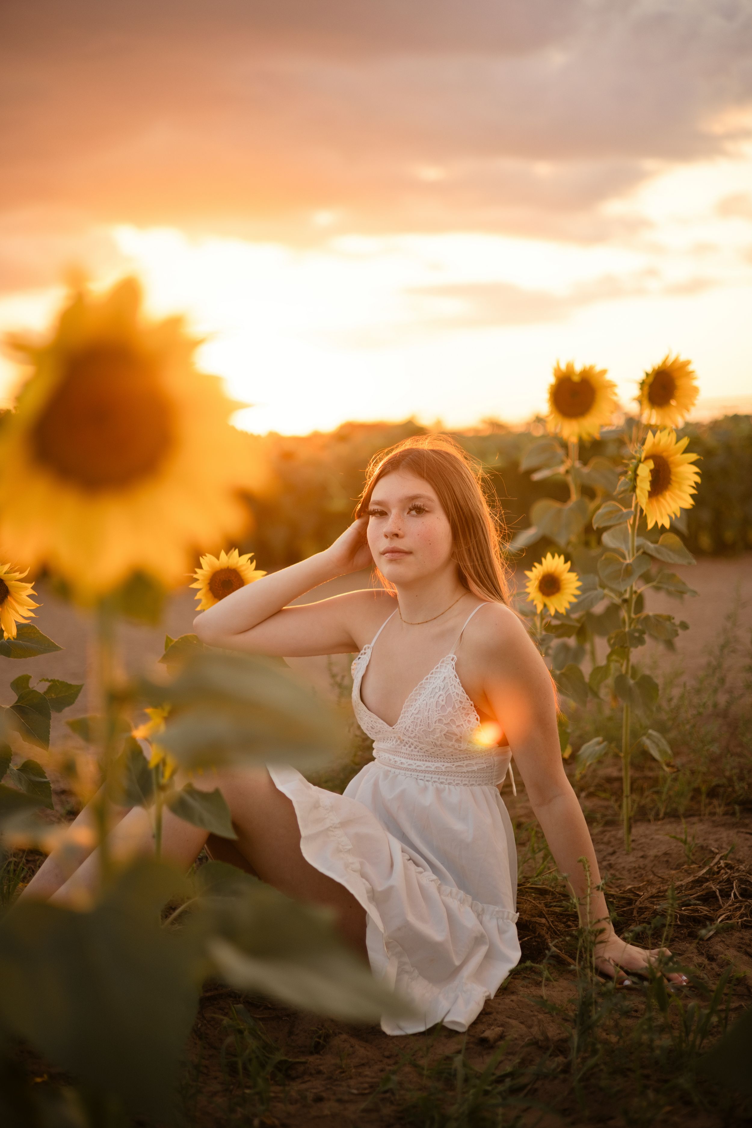 Young woman in white dress sitting in sunflower field at golden sunset, surrounded by blooming sunflowers.