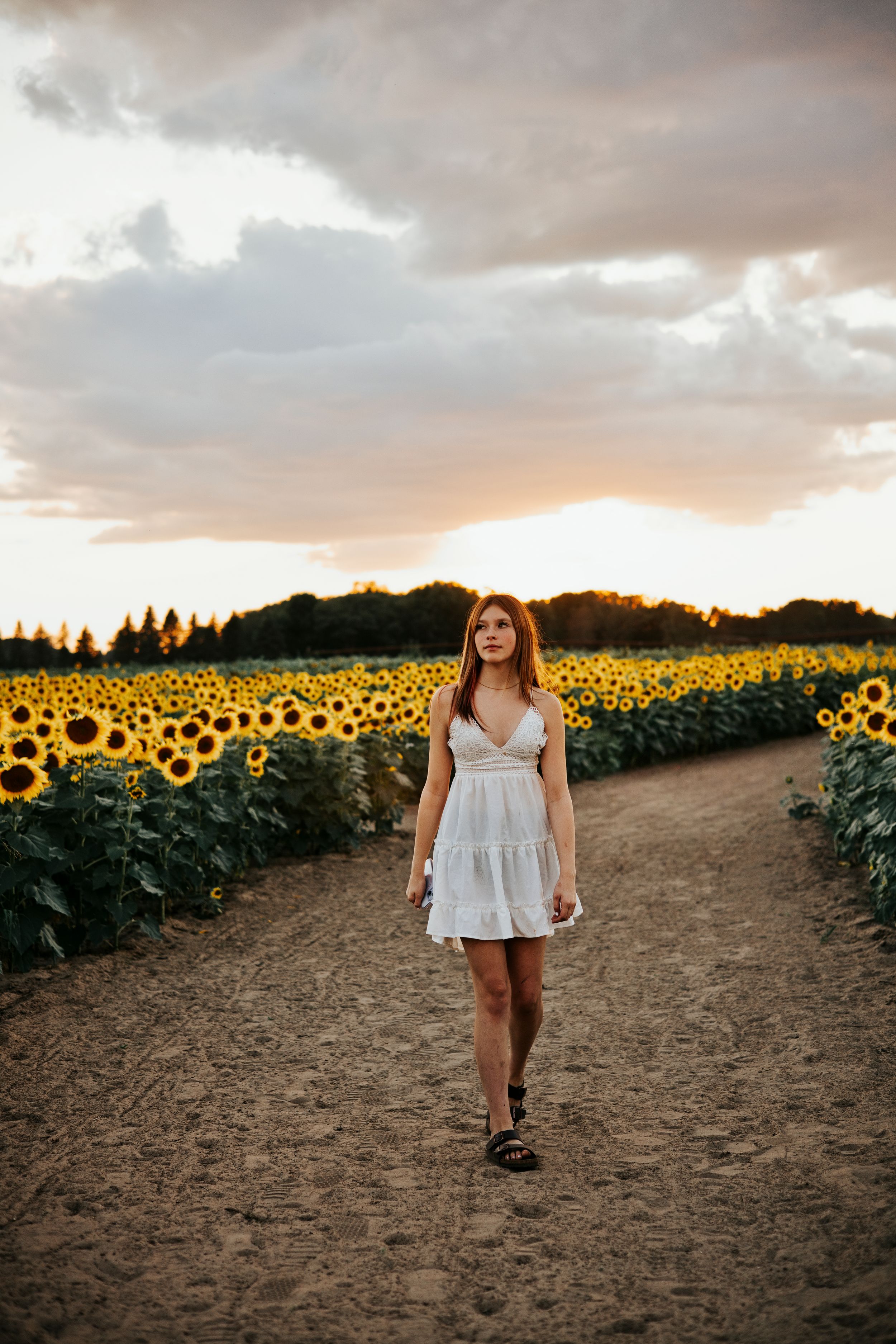 Young woman in white dress walking through a sunflower field at sunset with dramatic cloudy sky.