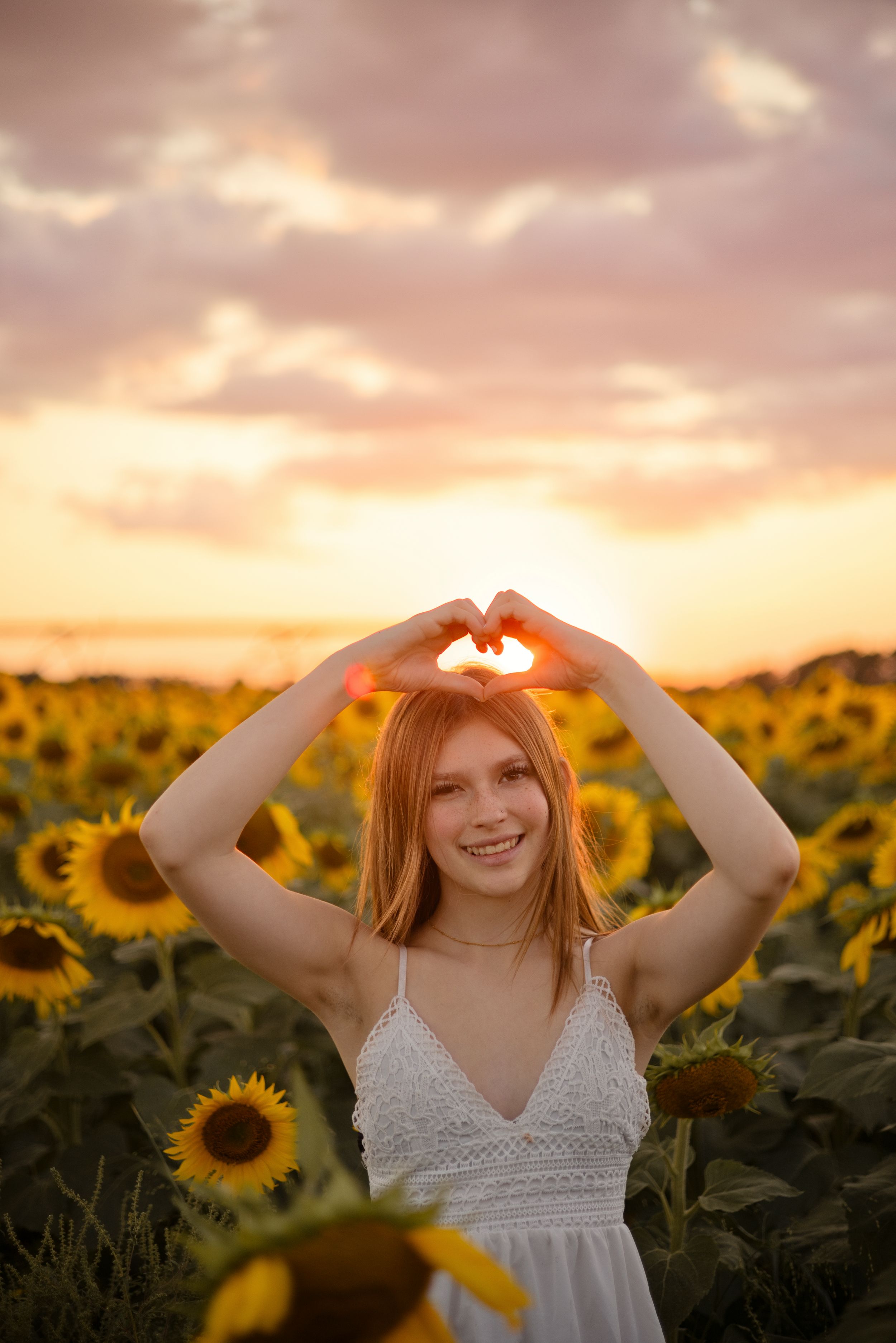 Young redhead woman in white dress making heart shape with hands in sunflower field at golden sunset.