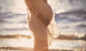 Pregnant woman in lace dress standing on beach with golden bokeh sunlight reflecting off ocean water.