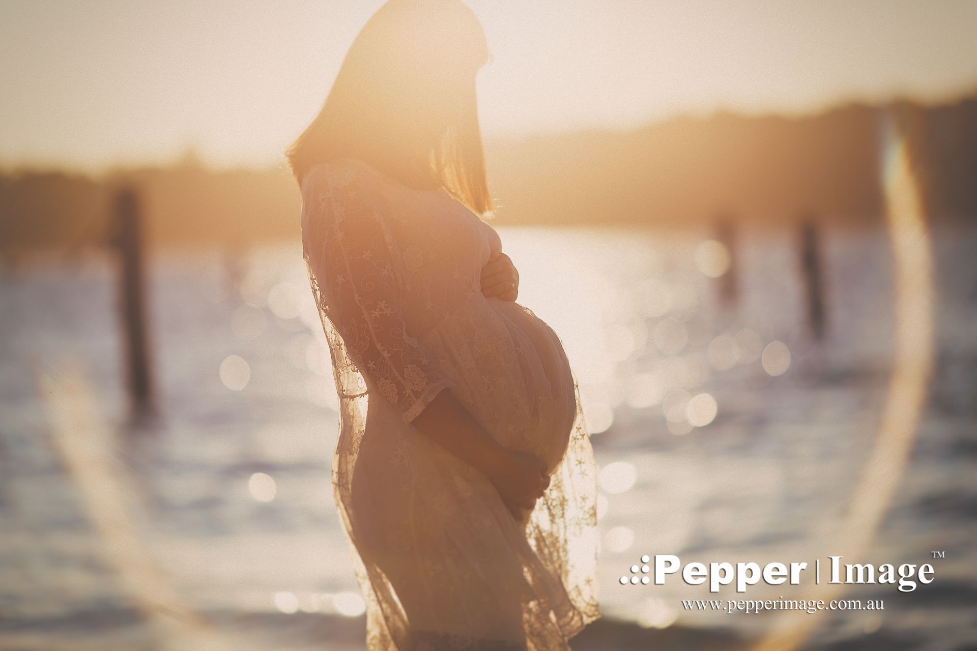 Pregnant woman standing in golden sunset light by the water, silhouetted in a flowing sheer dress.