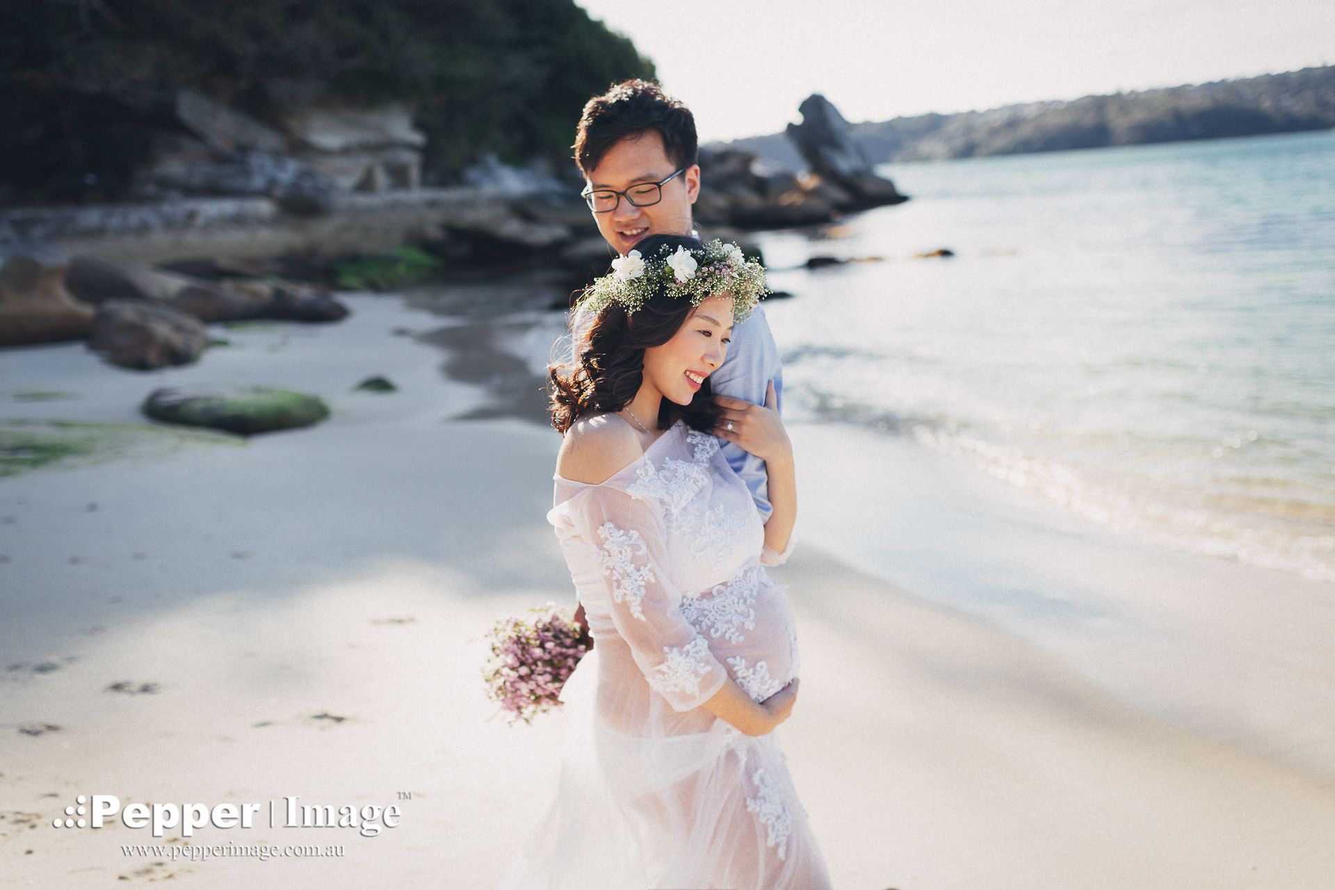 Couple posing on a sandy beach, woman in white lace dress holding bouquet, man embracing her, ocean in background.
