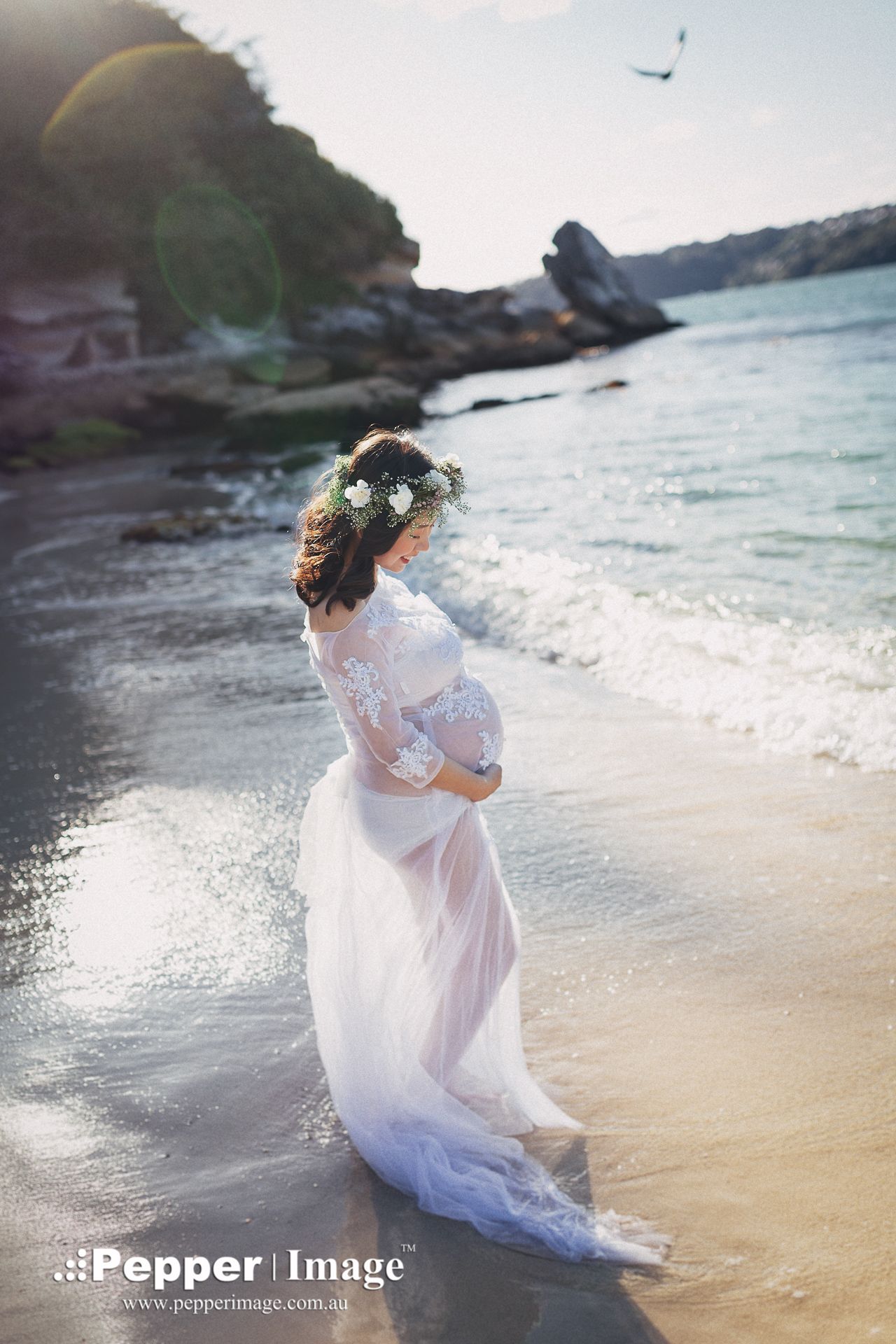 Pregnant woman in white lace gown and floral crown stands on a sunlit beach with ocean waves at her feet.