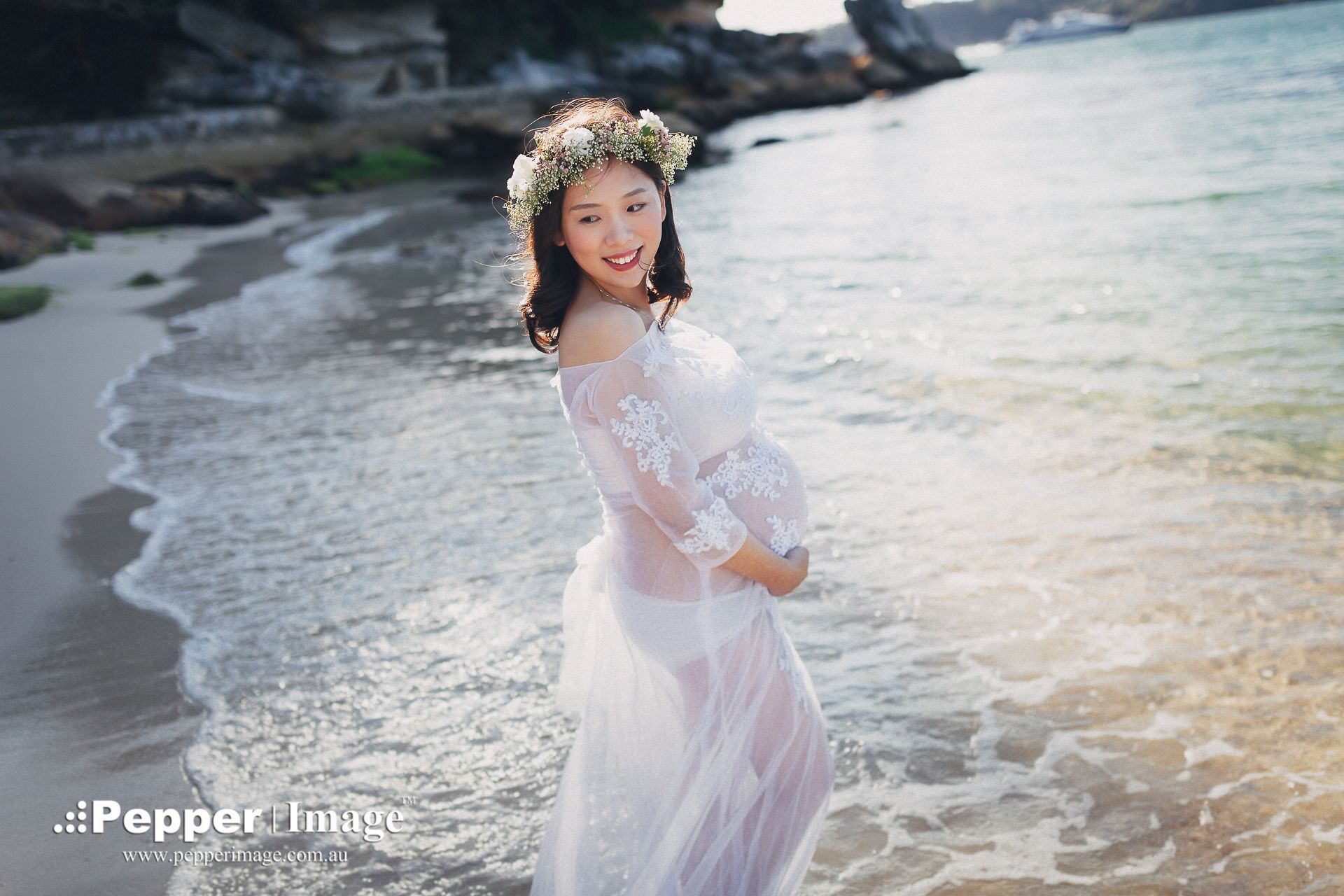 A smiling woman in a white lace dress and floral crown poses gracefully on a sandy beach with gentle ocean waves.