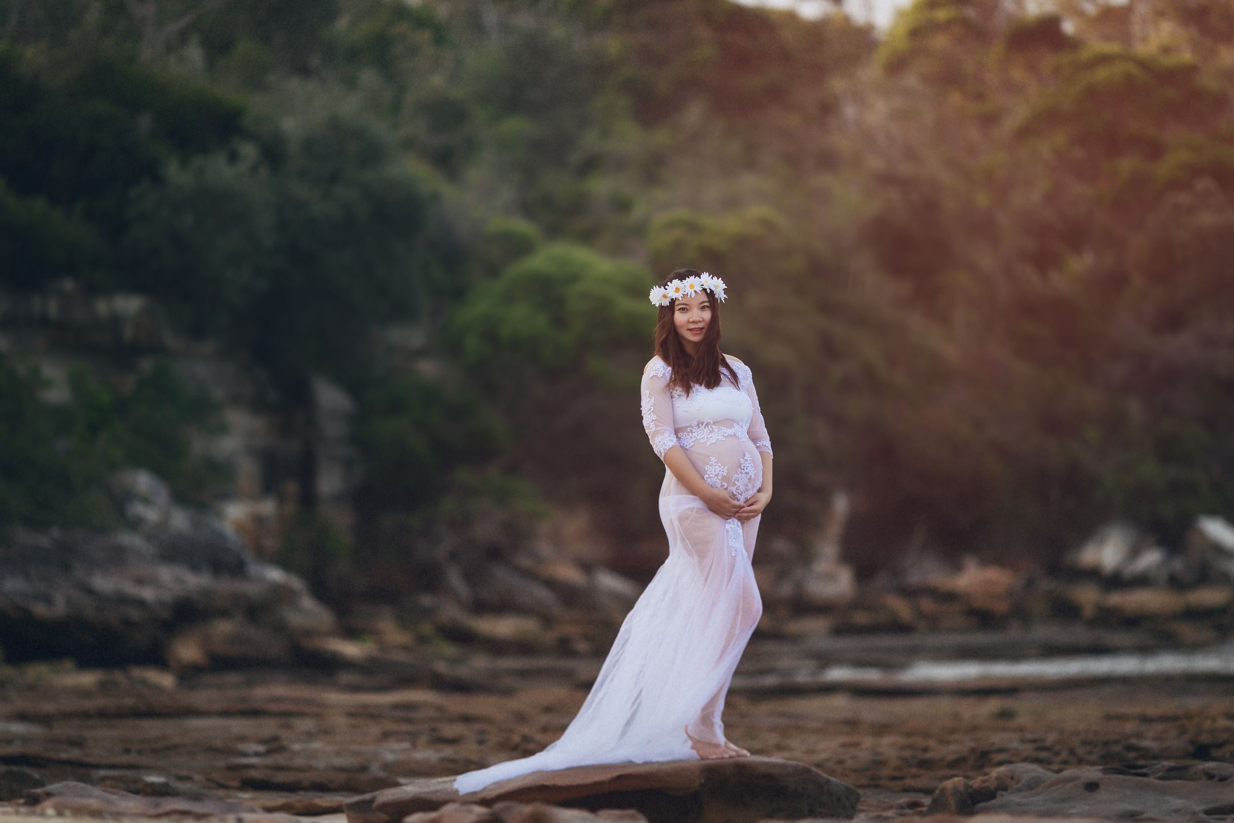 Pregnant woman in white lace gown and floral crown poses on rocky beach surrounded by lush greenery.