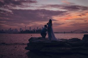 Couple in wedding attire silhouetted against a vibrant purple sunset over Sydney Harbour skyline on rocky shore.
