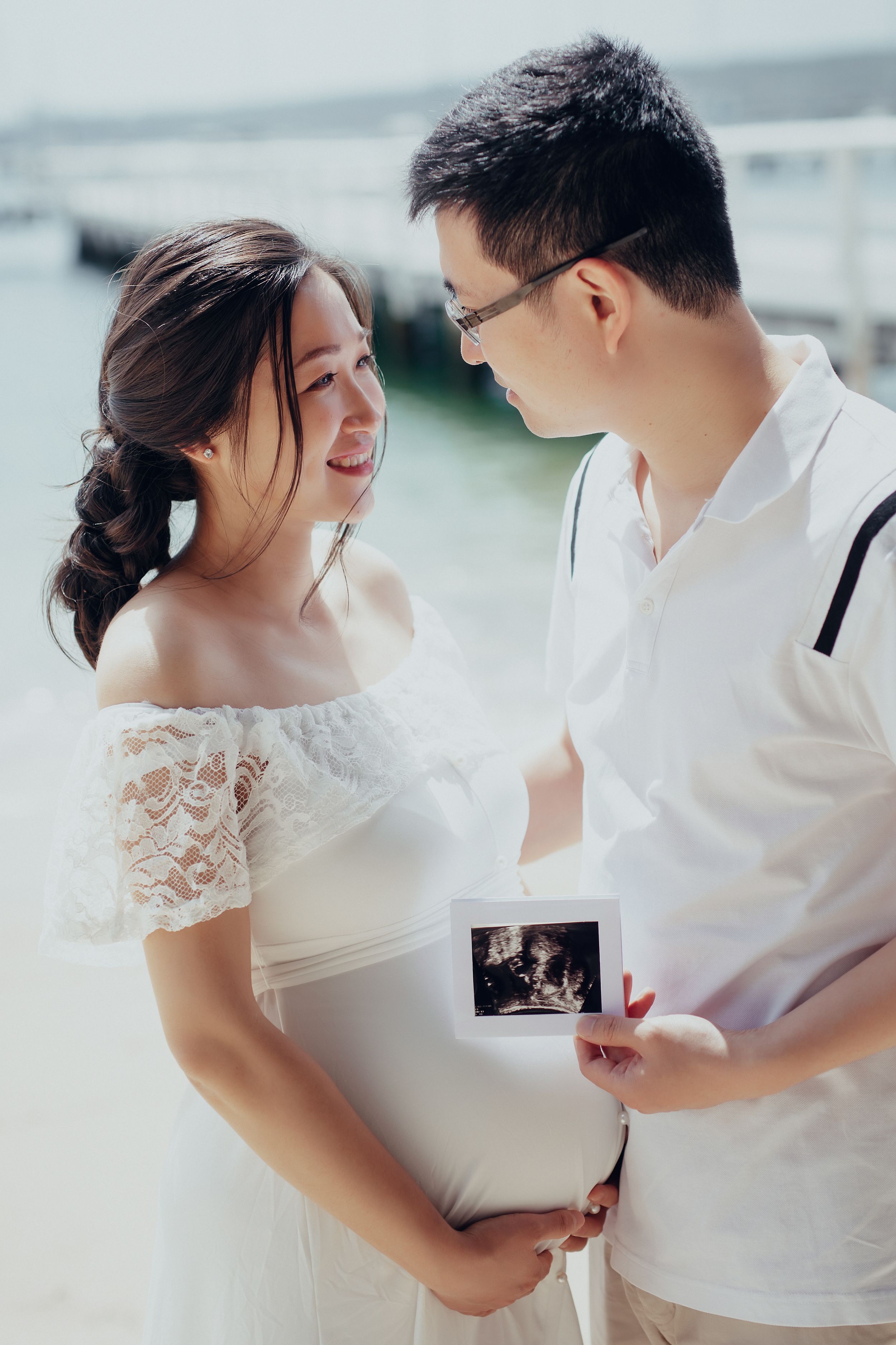 Smiling pregnant Asian couple holding ultrasound photo outdoors near water, woman in white lace dress, man in white shirt.