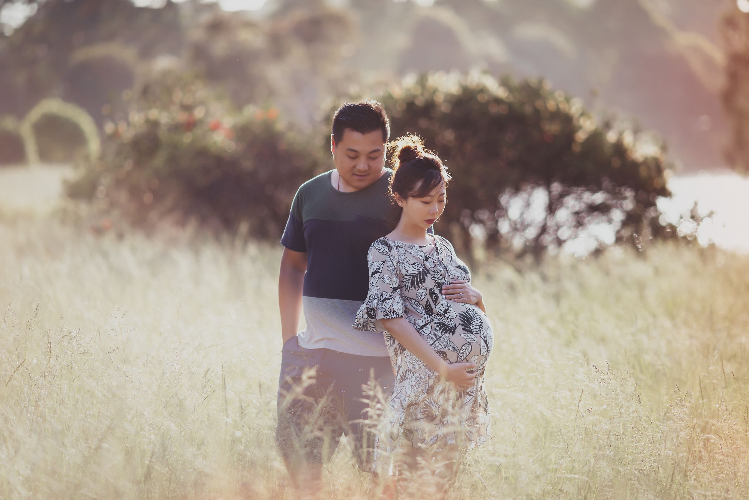 Pregnant couple standing in a sunlit field, man holding ultrasound photo while woman cradles her baby bump.