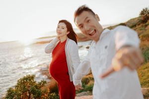 Pregnant woman in red dress poses with smiling man by the ocean at sunset.