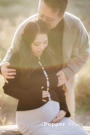 Couple sharing a tender maternity moment outdoors in warm golden light, man embracing pregnant woman from behind.