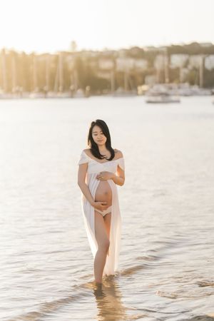 Pregnant woman in white sheer dress stands in shallow water at golden hour, cradling her baby bump near a marina.