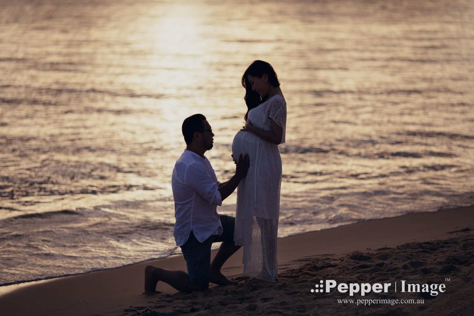 Silhouette of a man kneeling before a pregnant woman on a beach at golden sunset, romantic maternity photo.