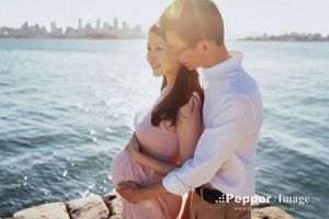 Pregnant couple embracing by the waterfront with a city skyline in the background during a maternity photo shoot.