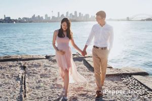 Pregnant couple holding hands walking along Sydney Harbour waterfront with city skyline in background.