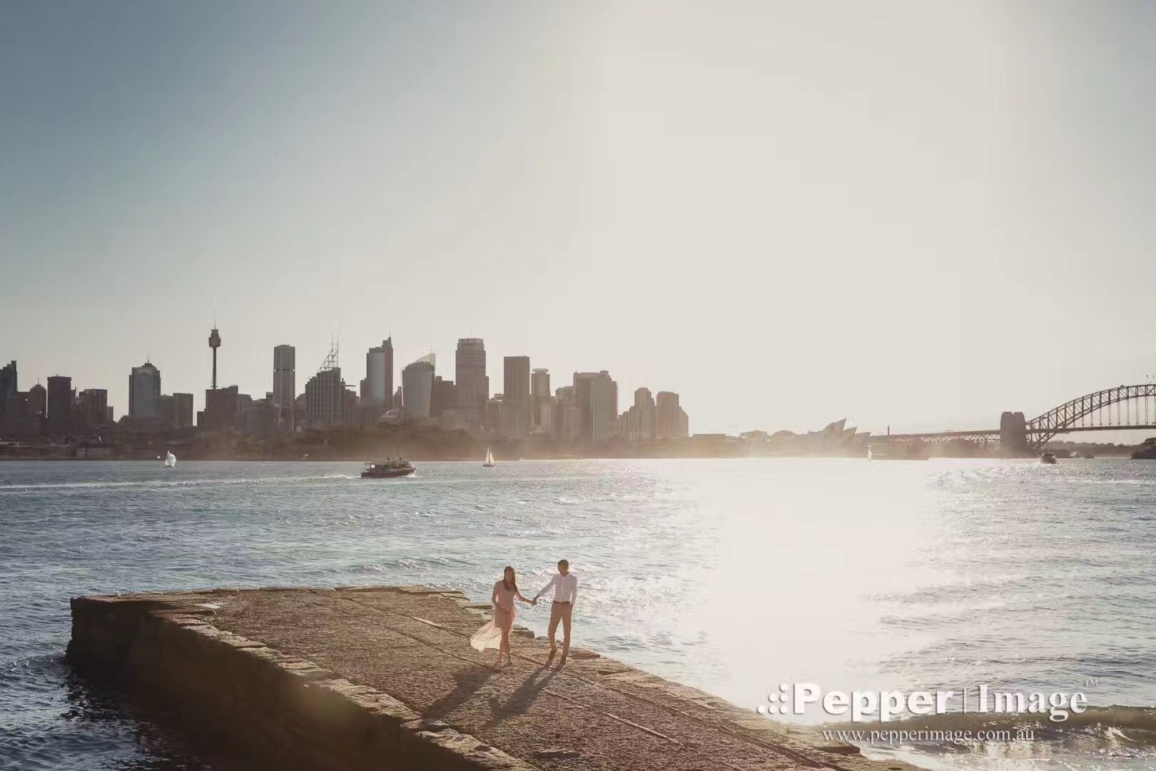 Couple holding hands on Sydney Harbour foreshore with iconic city skyline and Harbour Bridge at sunset.