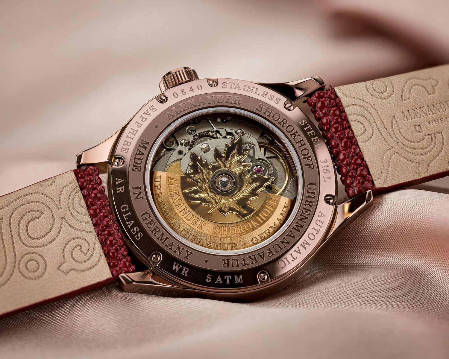 Rose gold skeleton watch with intricate engravings on case back, red strap, displayed on satin fabric.
