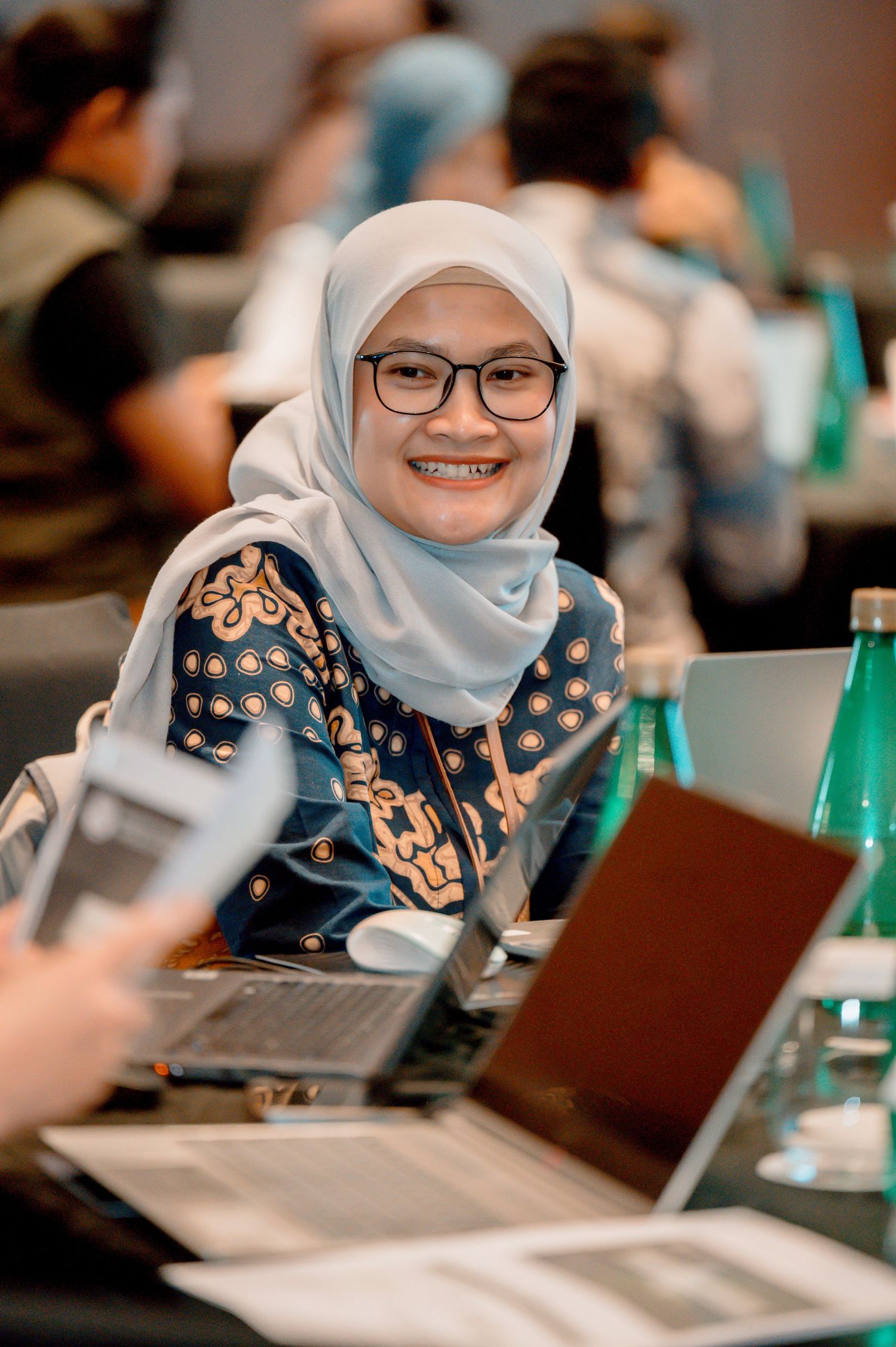 Smiling woman wearing hijab and glasses at conference table with laptops, dressed in batik top.