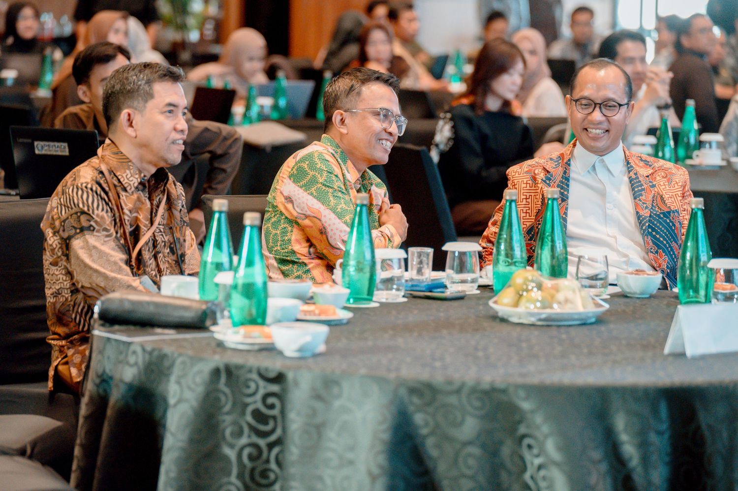 Three men in batik shirts converse at a formal conference table with green bottles and glasses in a banquet hall.