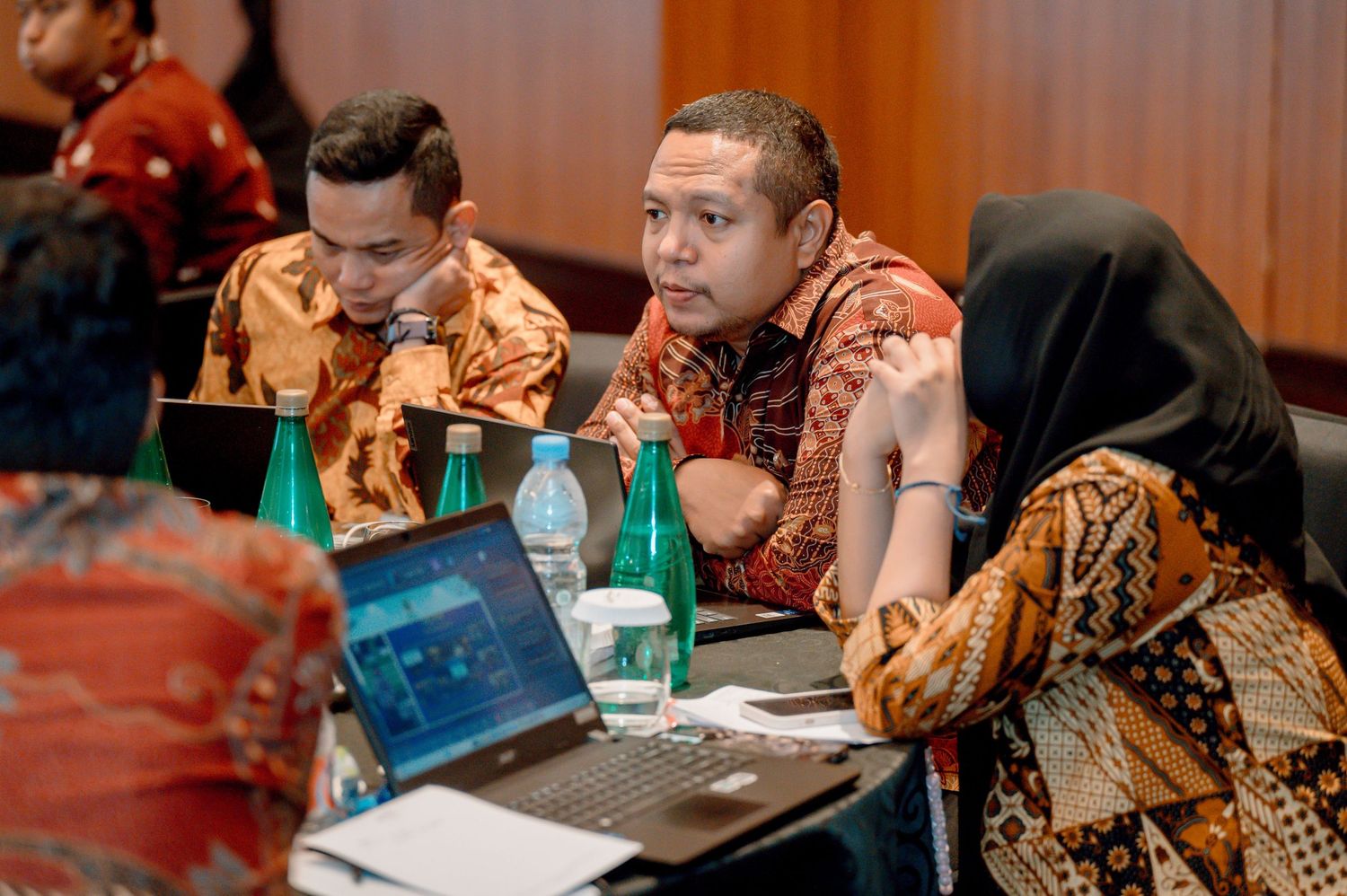 Professionals in batik shirts discuss work at a conference table with laptops and green water bottles.