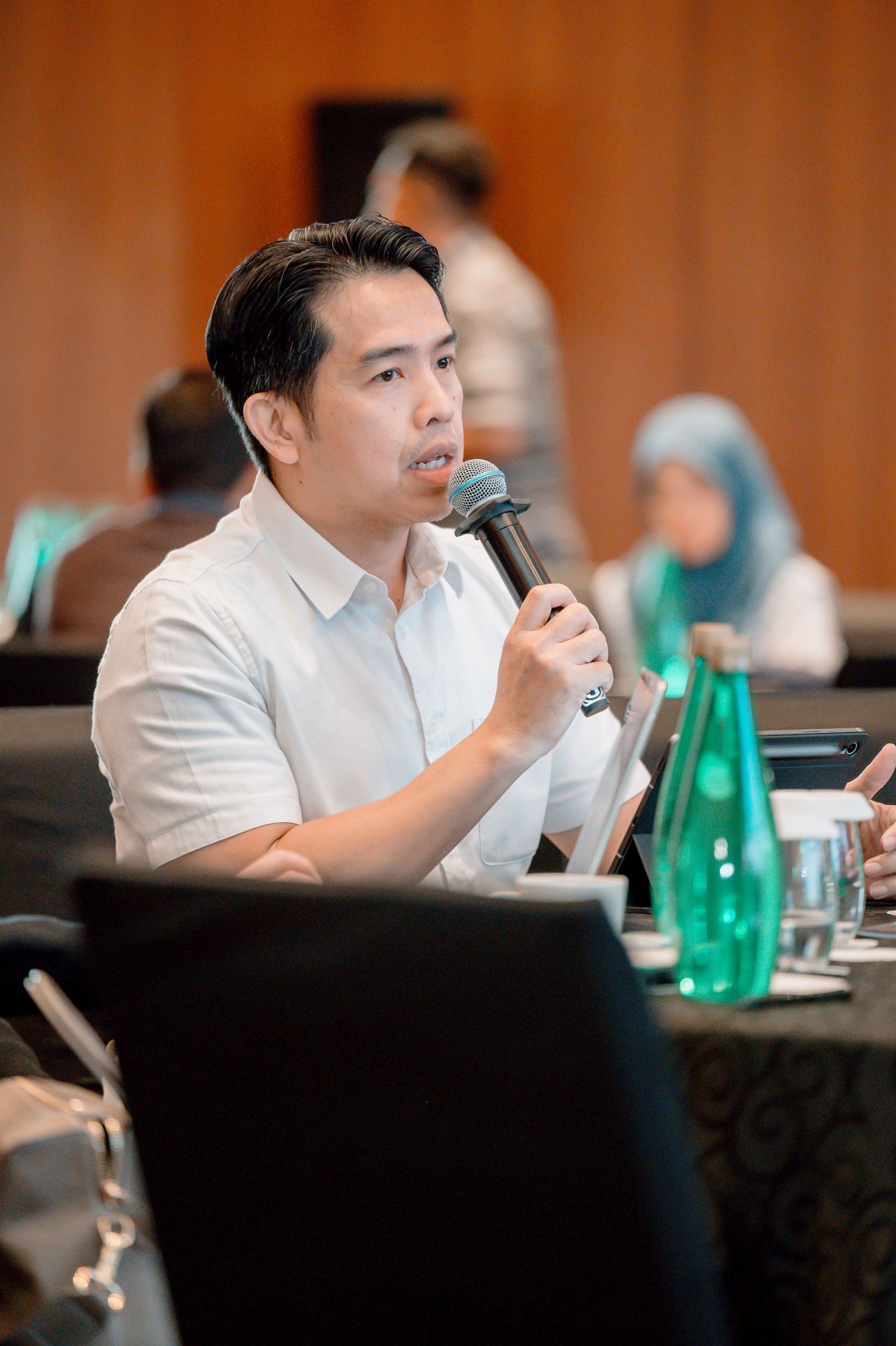 A man in a white shirt speaks into a microphone at a conference, holding a tablet, with attendees visible in the background.