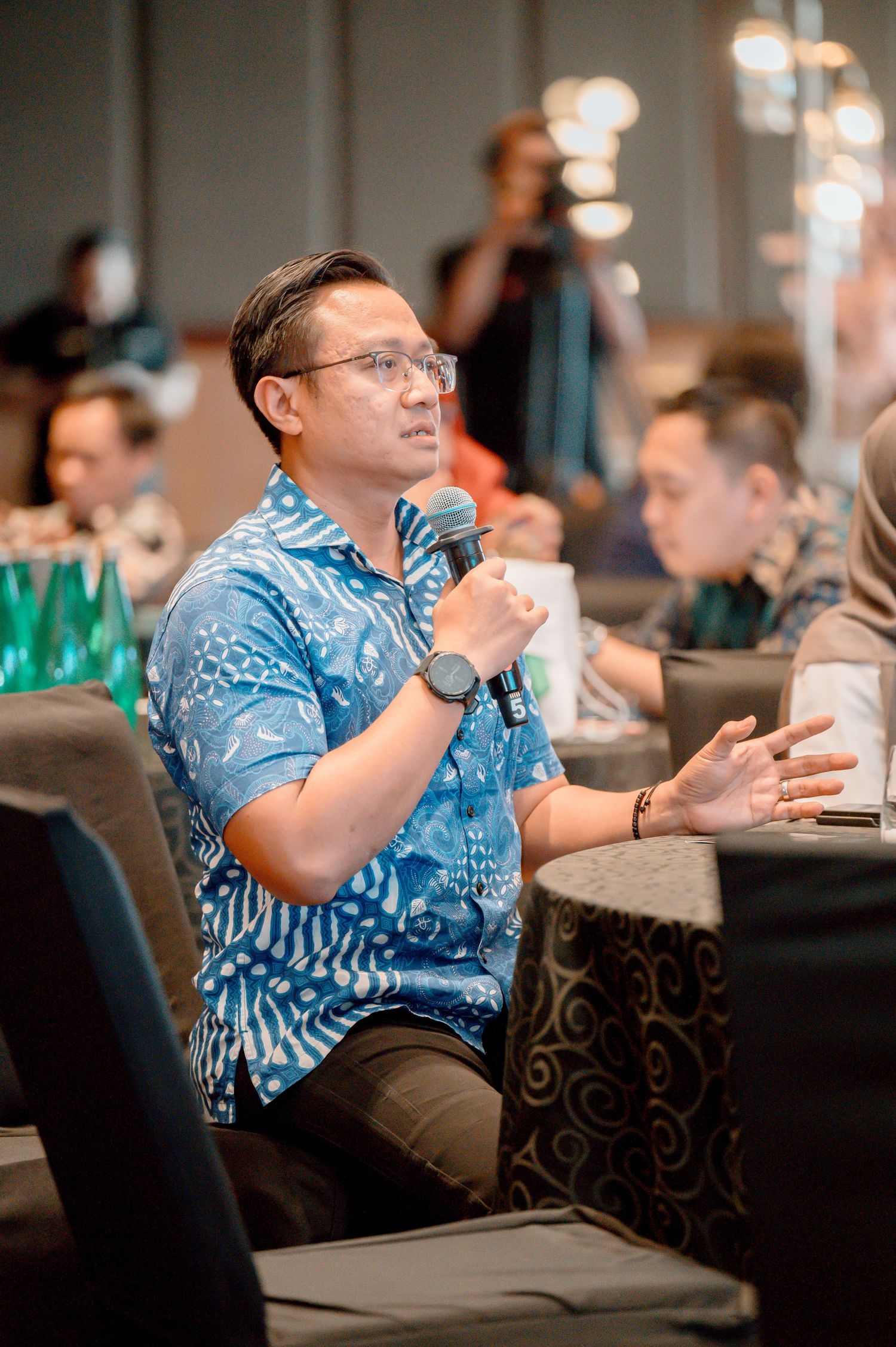 Man in blue batik shirt speaking at a conference panel, holding a microphone while gesturing expressively.