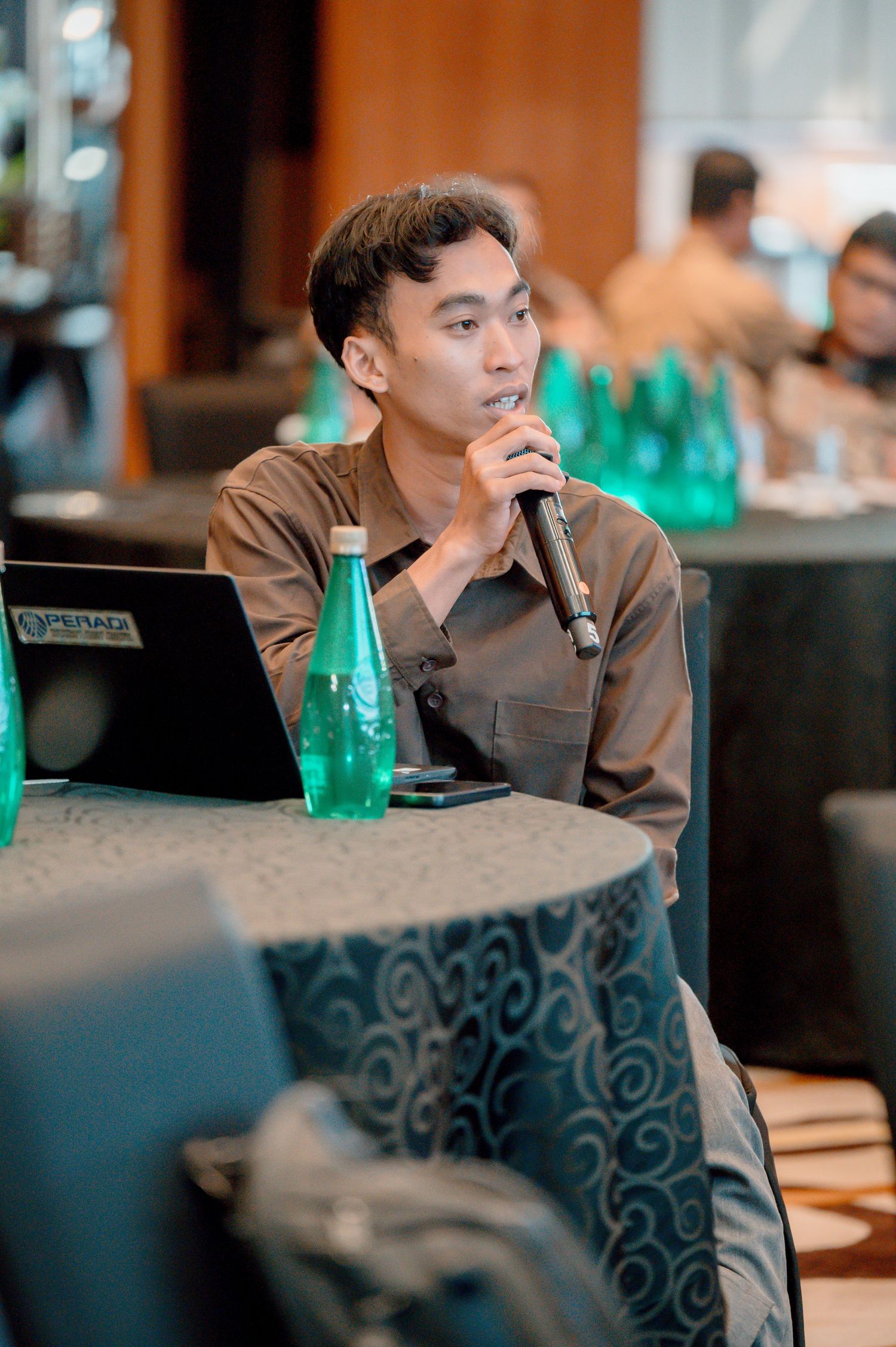 A young Asian man speaks into a microphone at a conference table with a laptop and green water bottles nearby.