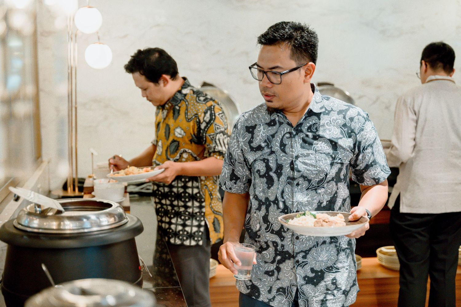 Men serving themselves at a buffet during a catered event, wearing batik shirts in a modern dining setting.