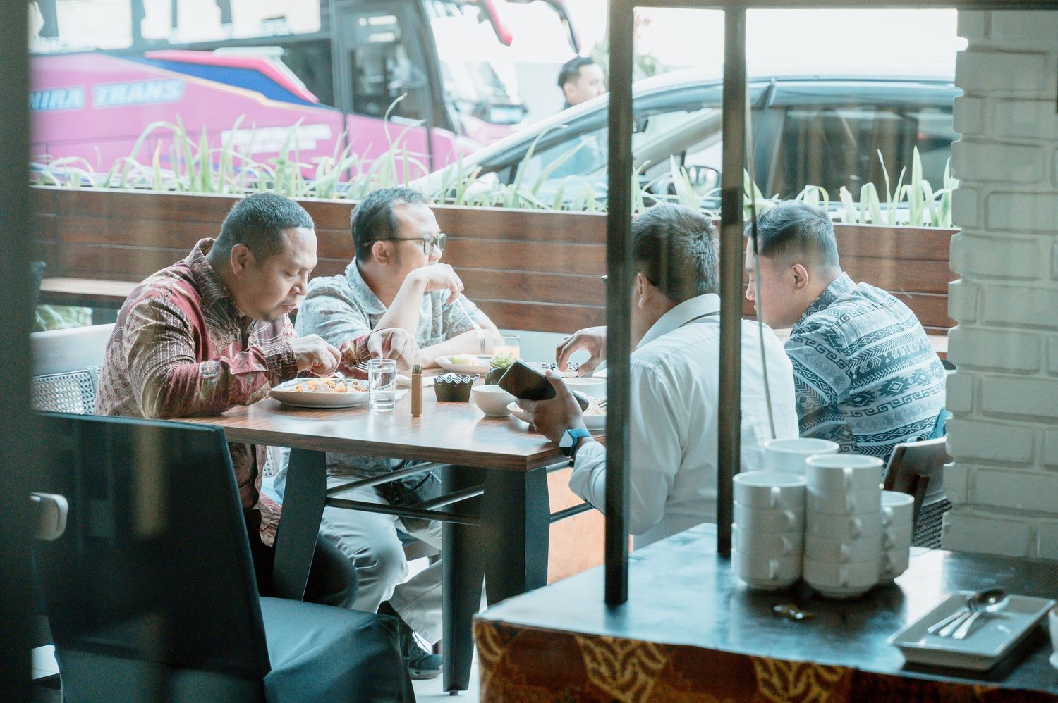 Men dining together at a restaurant table, viewed through a window, with cars visible outside.