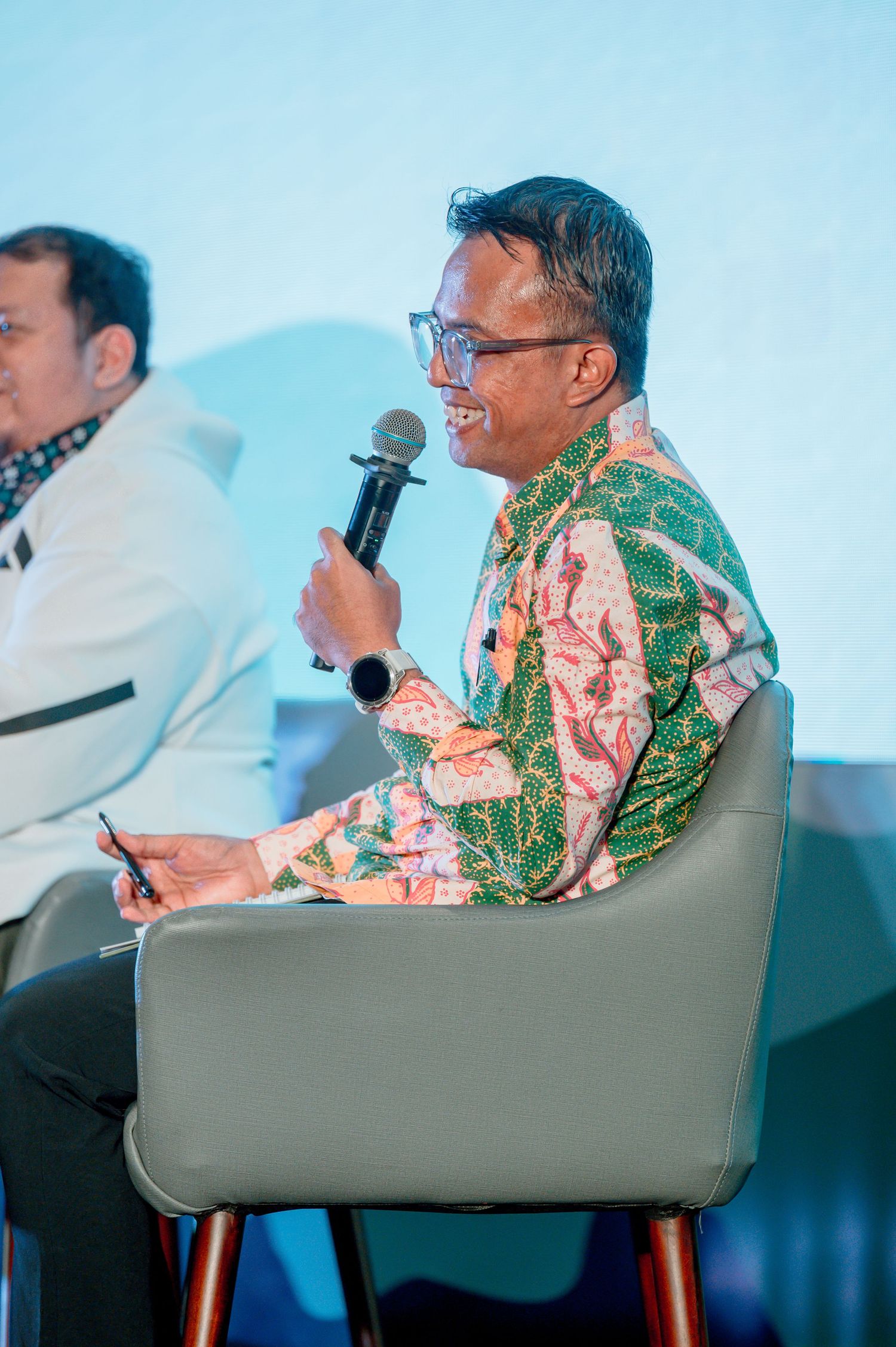 Man in batik shirt speaking into microphone while seated on stage at a conference panel discussion.