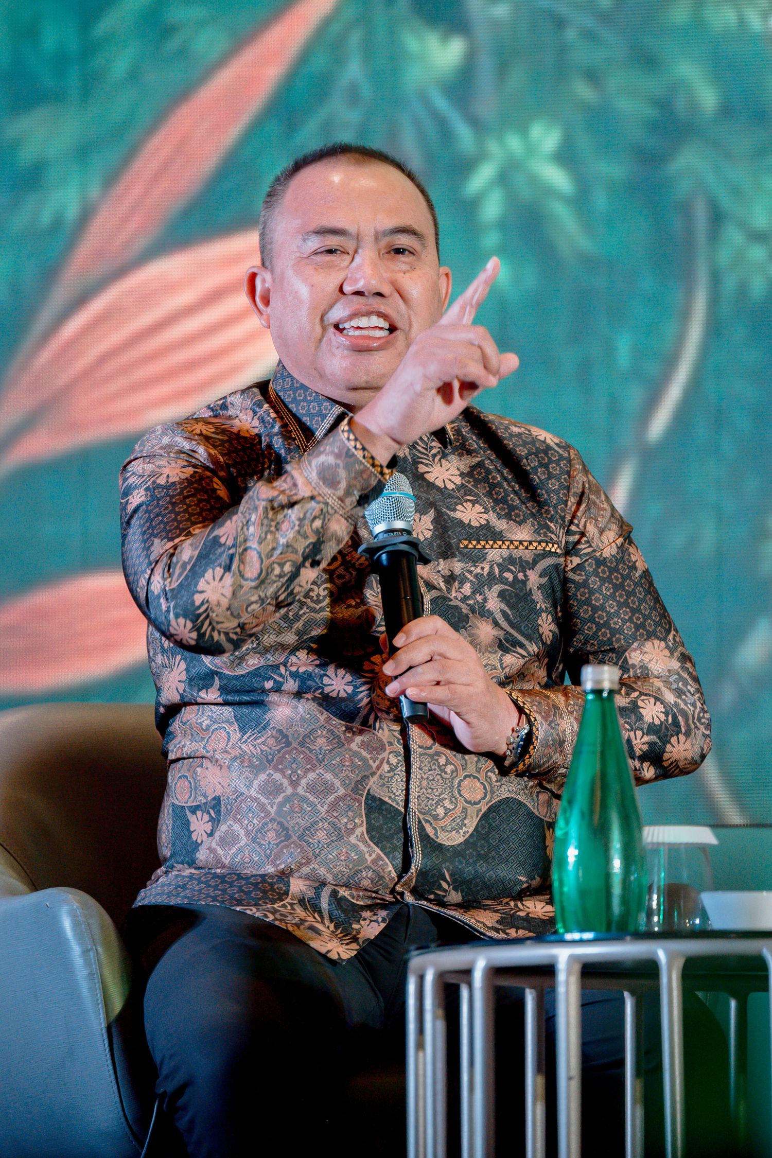 Man in batik shirt speaking at event, holding microphone and gesturing with hand, green botanical backdrop.