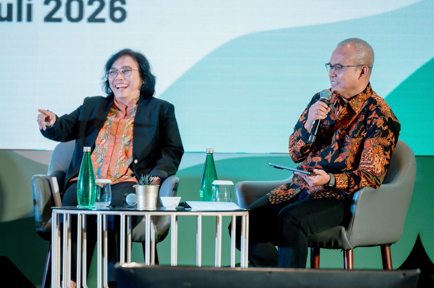 Two speakers engage in discussion on stage at a 2026 conference, one gesturing expressively while the other holds a microphone.