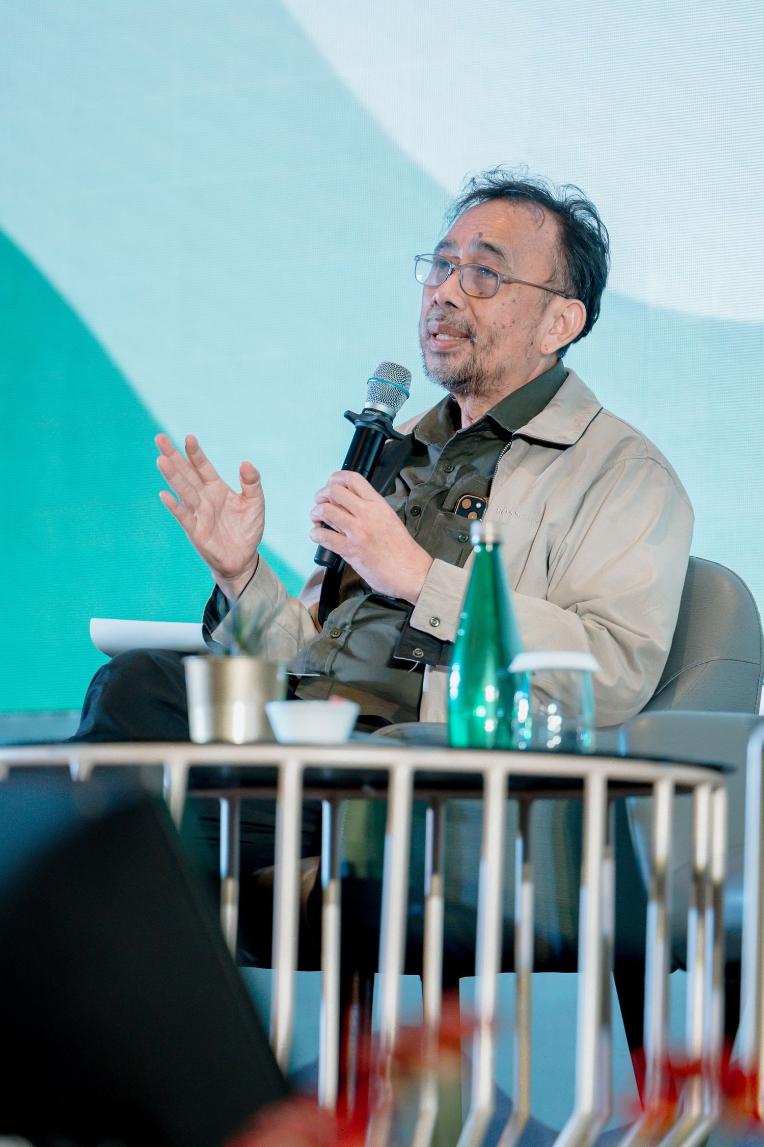 An elderly man speaks into a microphone at a panel discussion, gesturing with one hand against a teal backdrop.