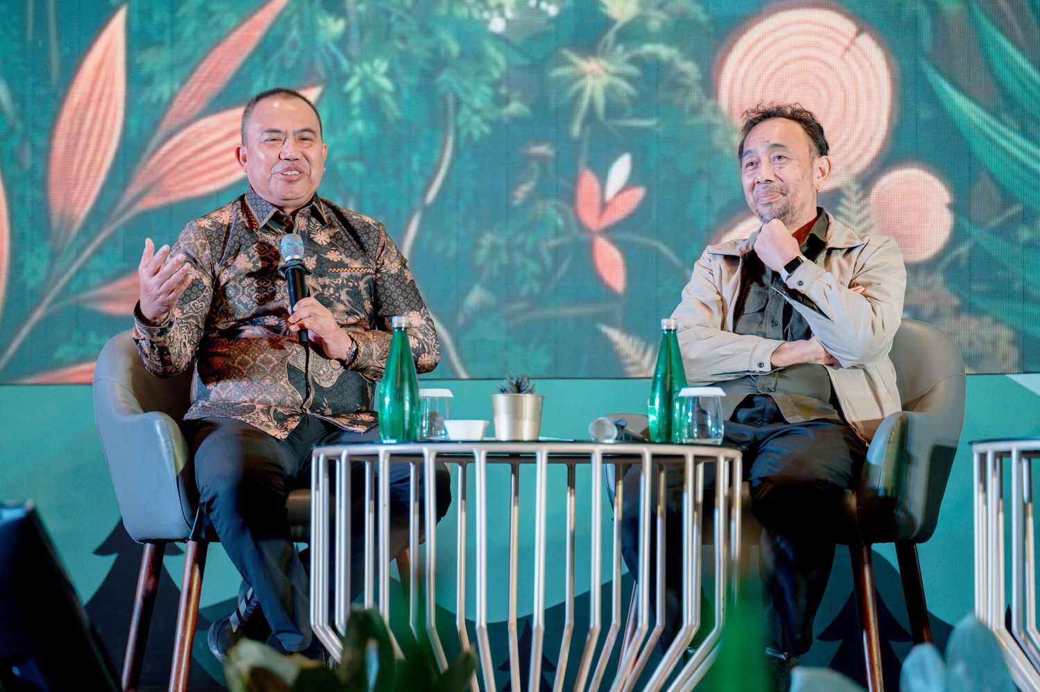 Two men in batik shirts engaged in discussion on stage with tropical green backdrop at a formal event.