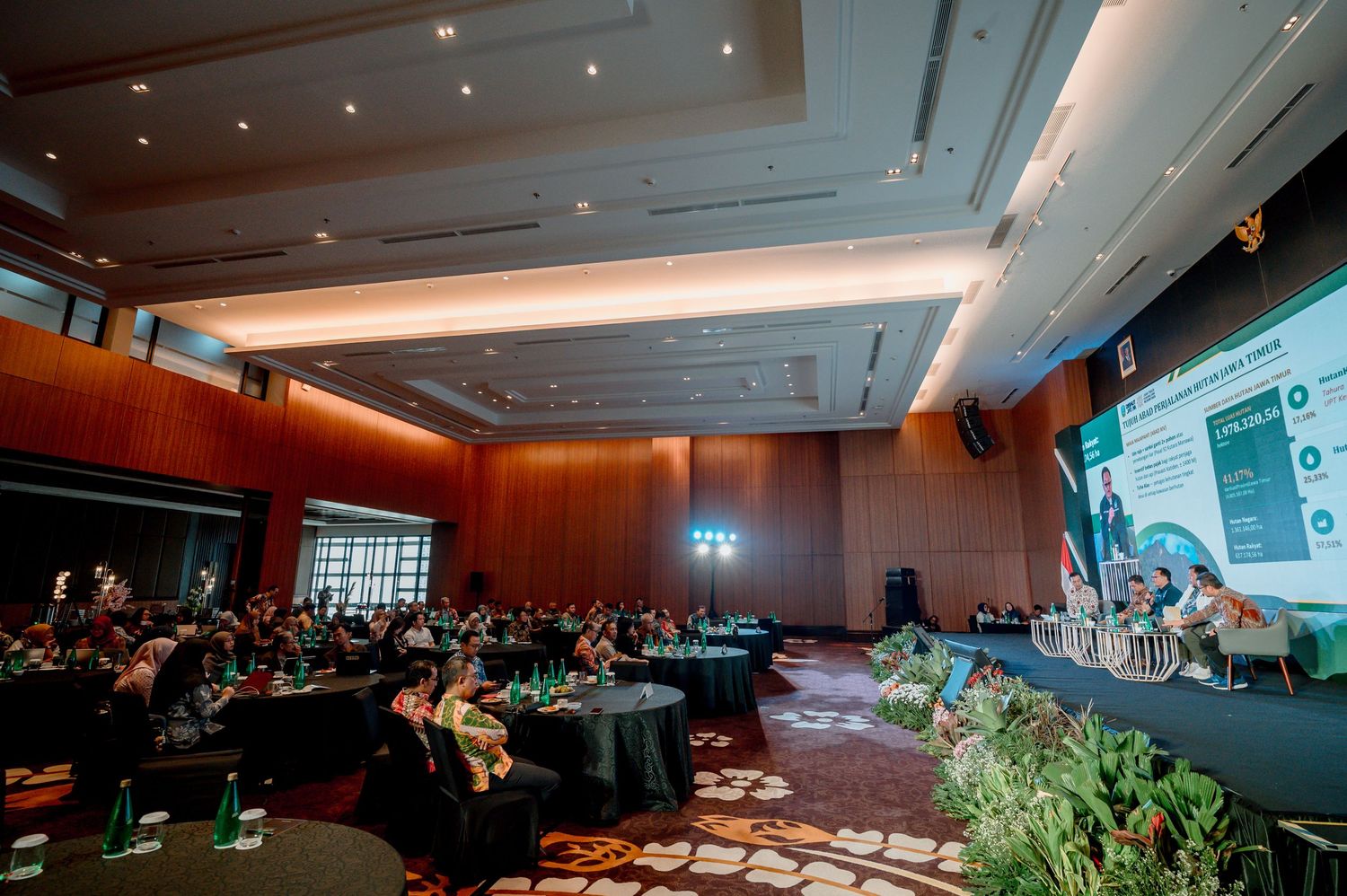 Large conference hall with audience seated at round tables facing a presentation screen and panel of speakers.