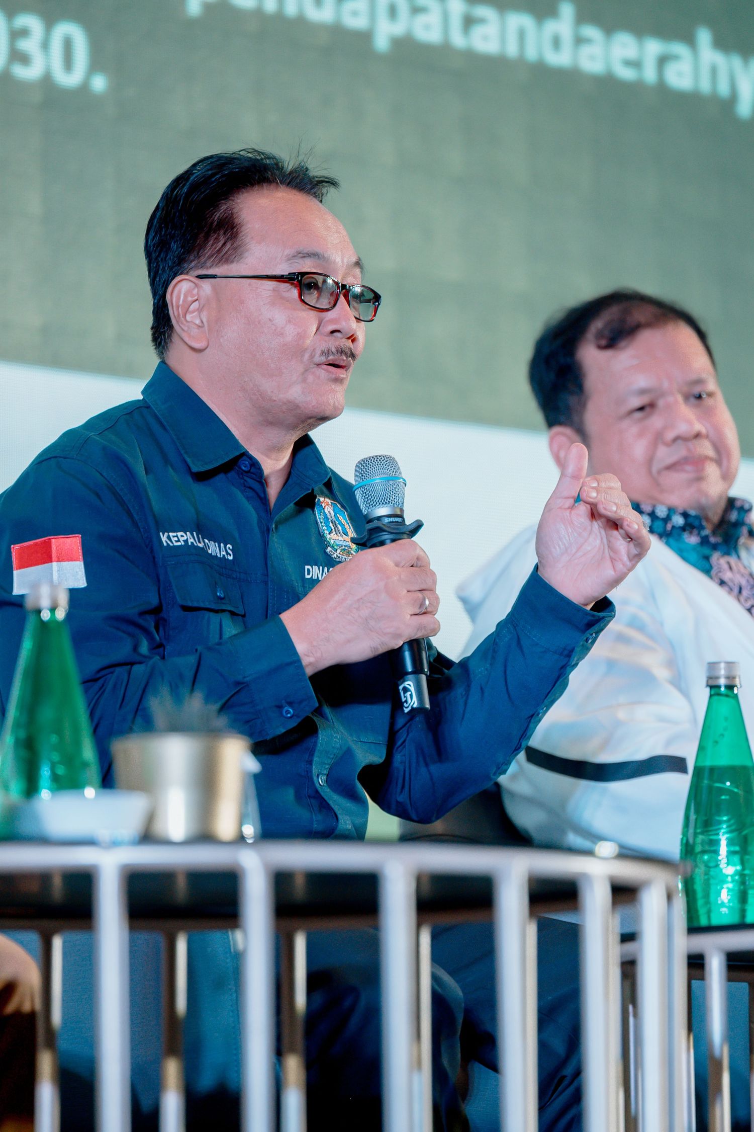 Indonesian official in blue uniform speaks at panel discussion, holding microphone with colleague seated beside him.