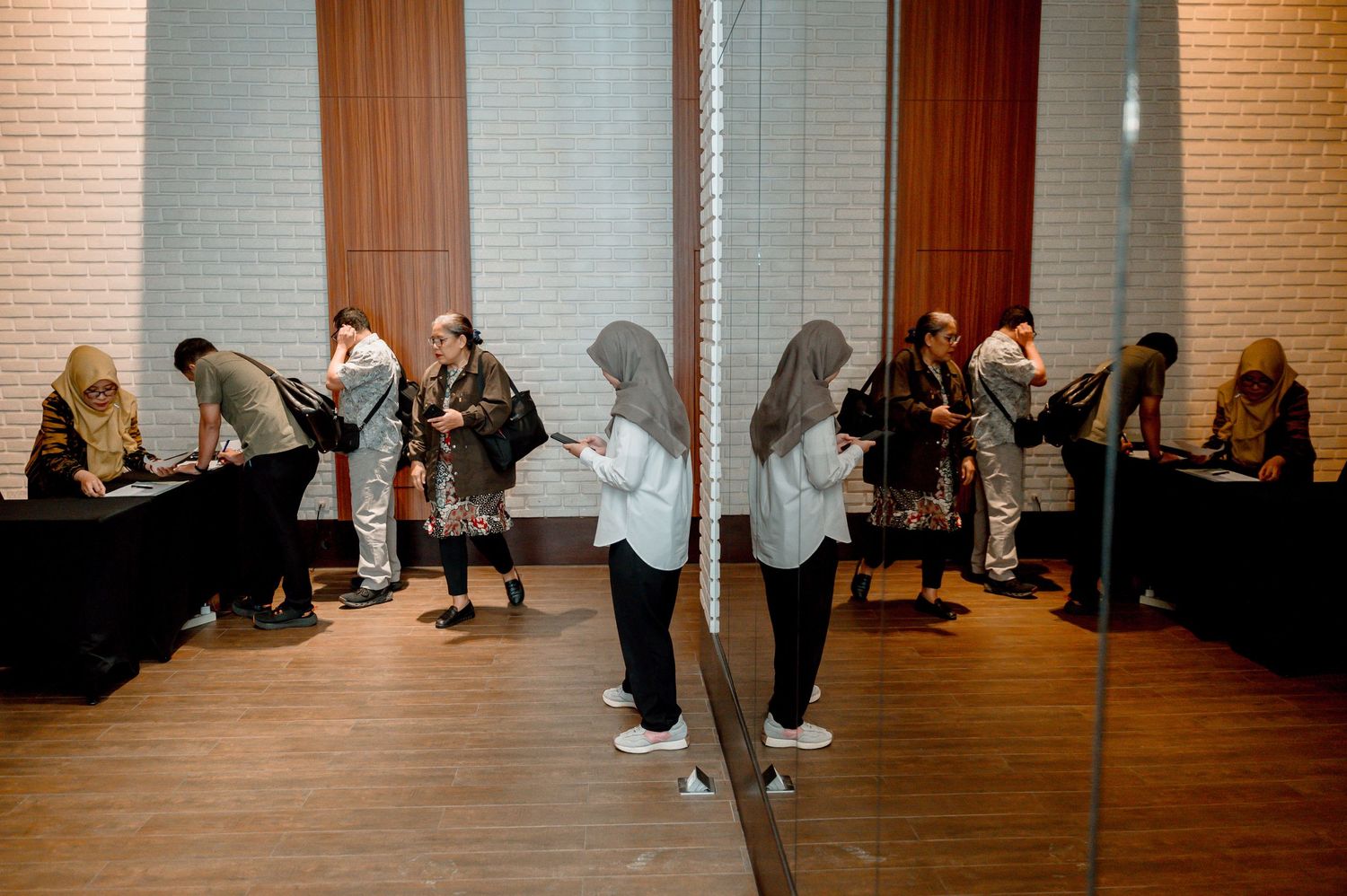 Women in hijabs check phones and sign in at a registration table in a modern hall, reflected in a mirror.