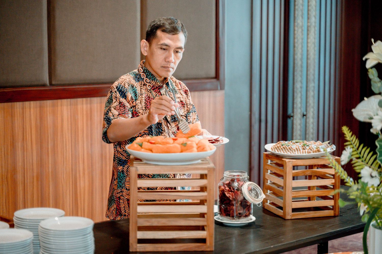 Man in floral shirt serving food at a buffet table with wooden crates holding dishes and snacks.