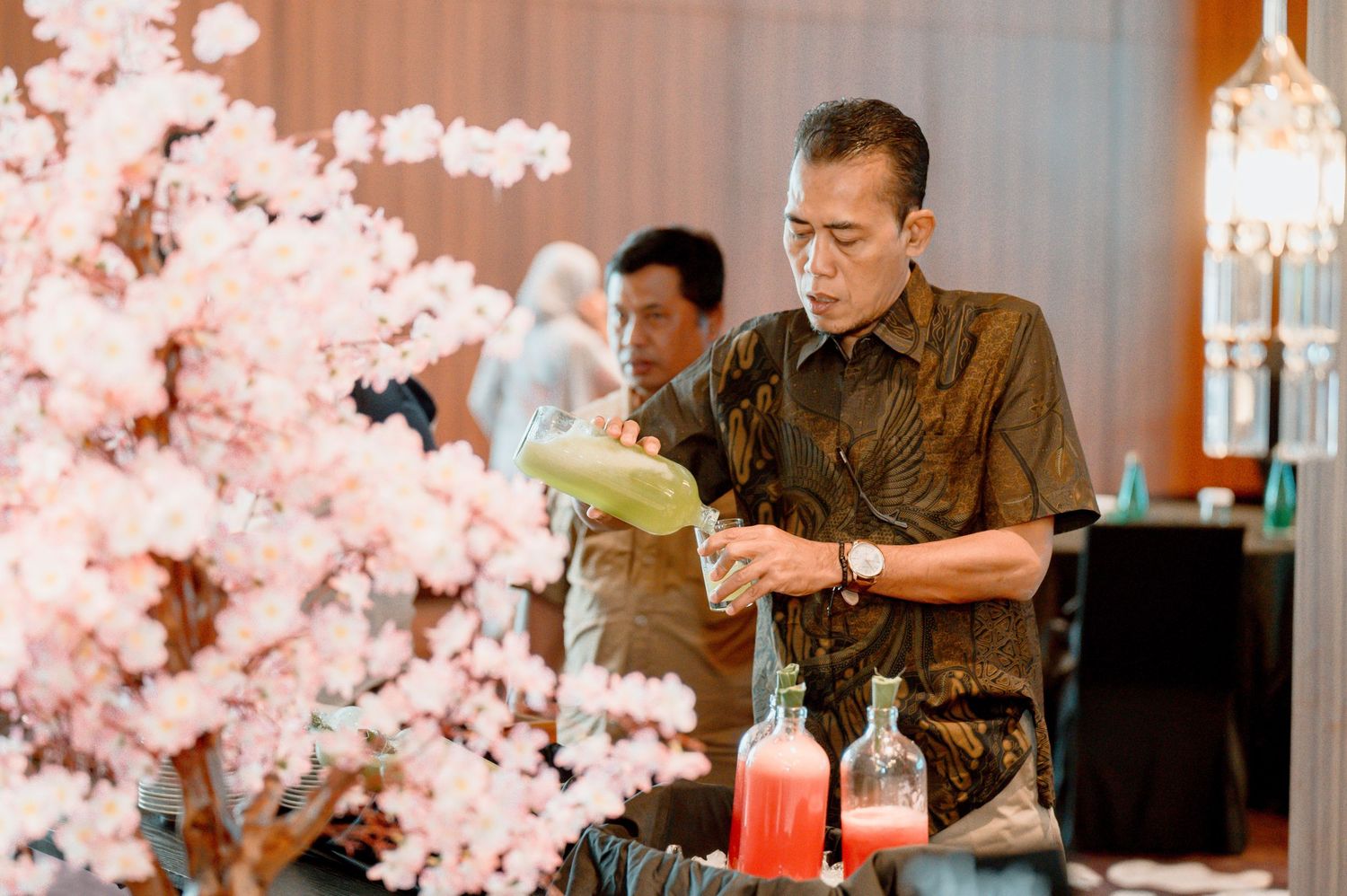 Man in batik shirt pouring drink at elegant event with pink cherry blossom decorations and red bottles.
